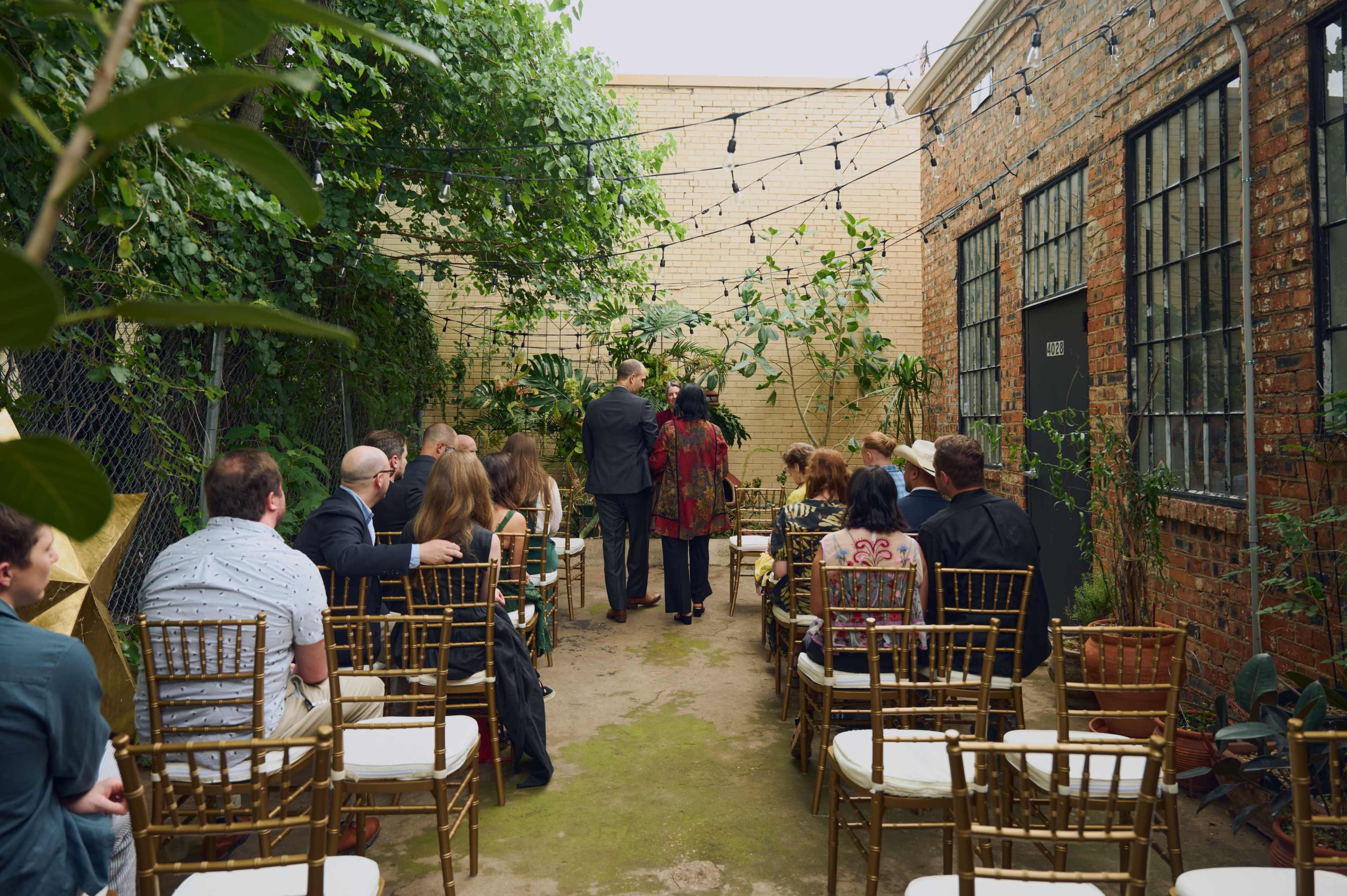 A group of people is seated in chairs facing an altar in a decorative outdoor space surrounded by plants and string lights.
