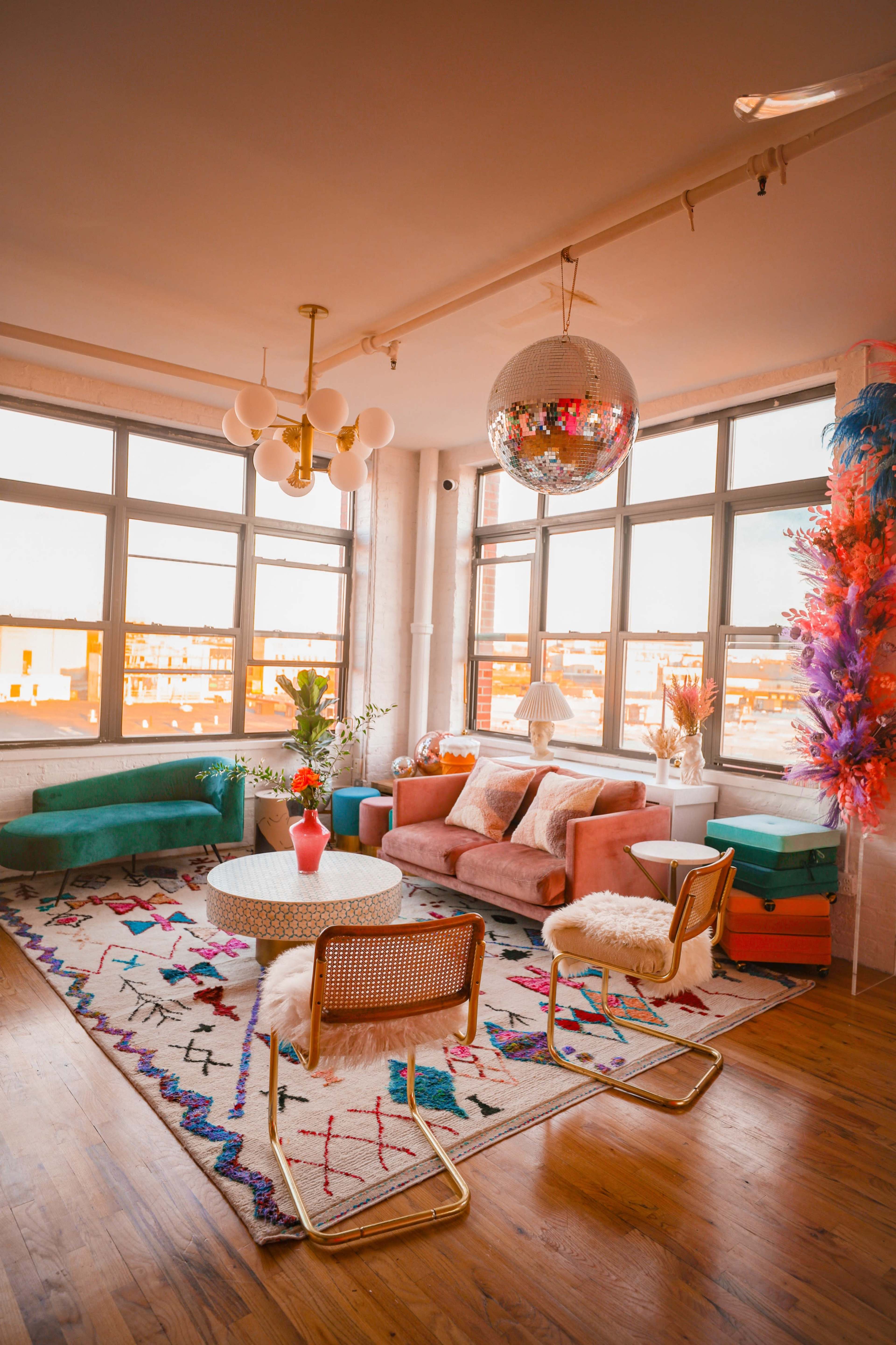 A colorful living room features large windows, a circular coffee table, and various seating options, including a pink sofa and green chairs, on a patterned rug.