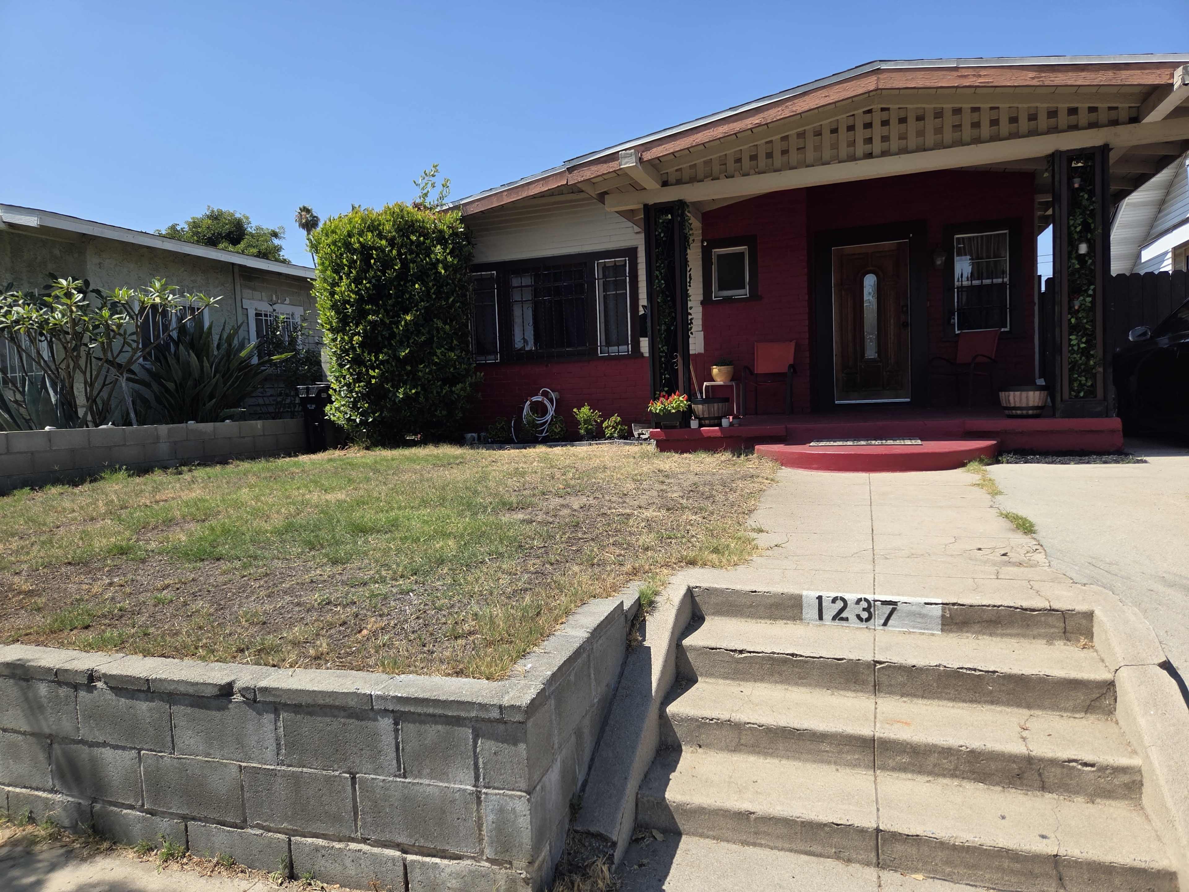 The image shows a single-story red house with a front porch, surrounded by a low gray wall and featuring a small lawn.