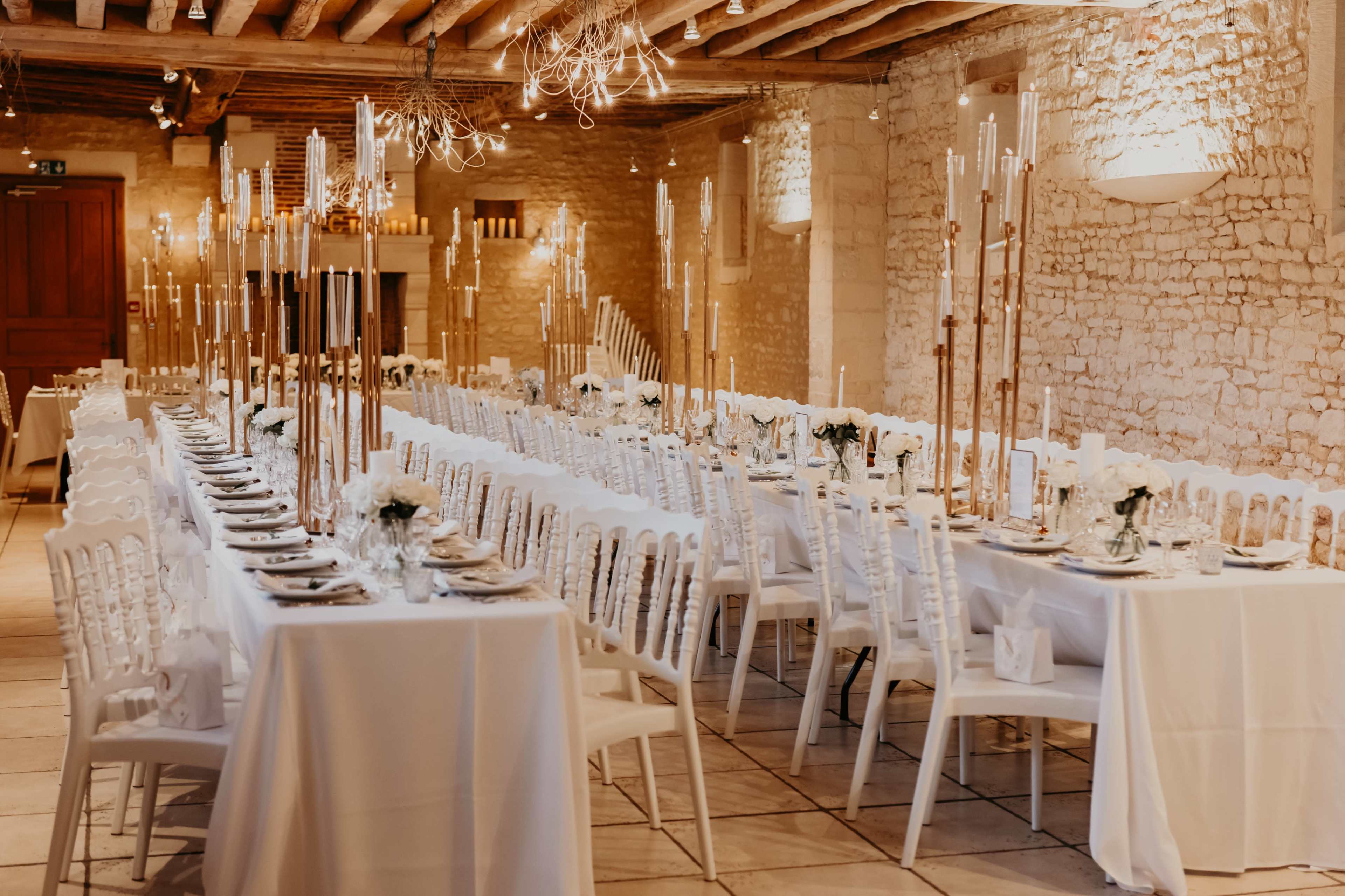 The image shows a dining hall set for a formal event, featuring long tables covered in white tablecloths and adorned with tall candle holders.