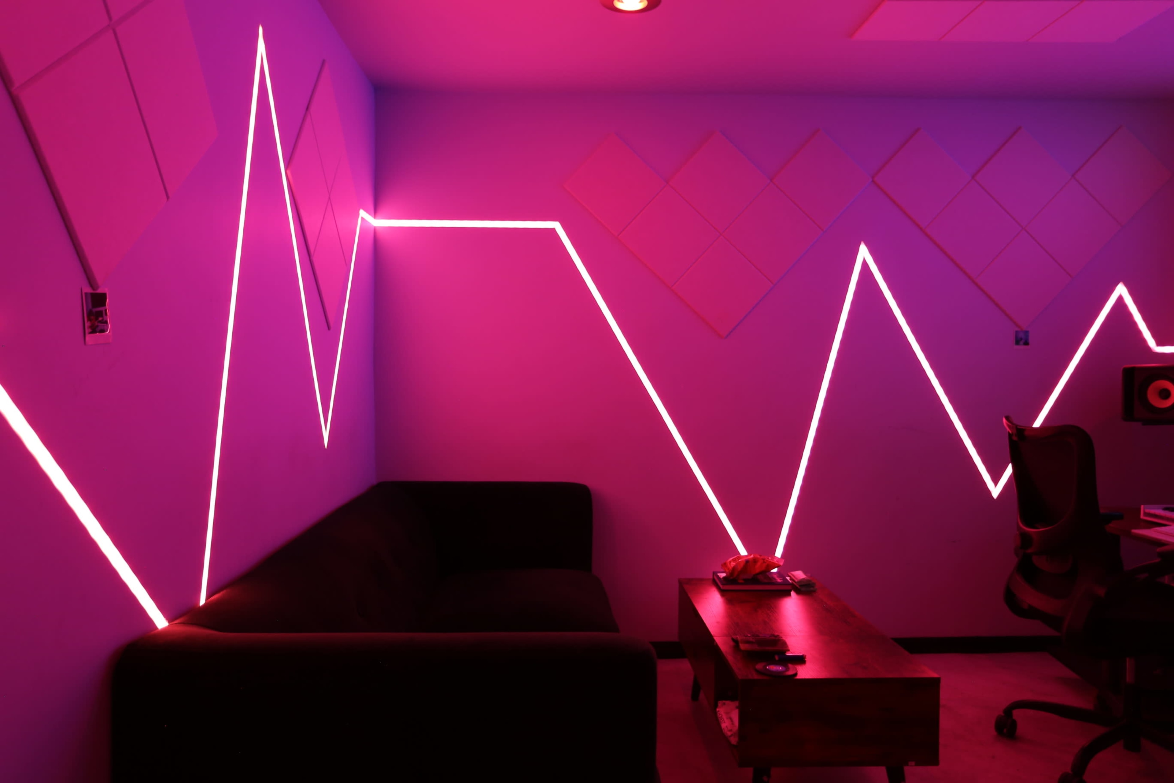 A room features a dark couch, a wooden table, and geometric pink neon lights outlining the walls in a zigzag pattern.