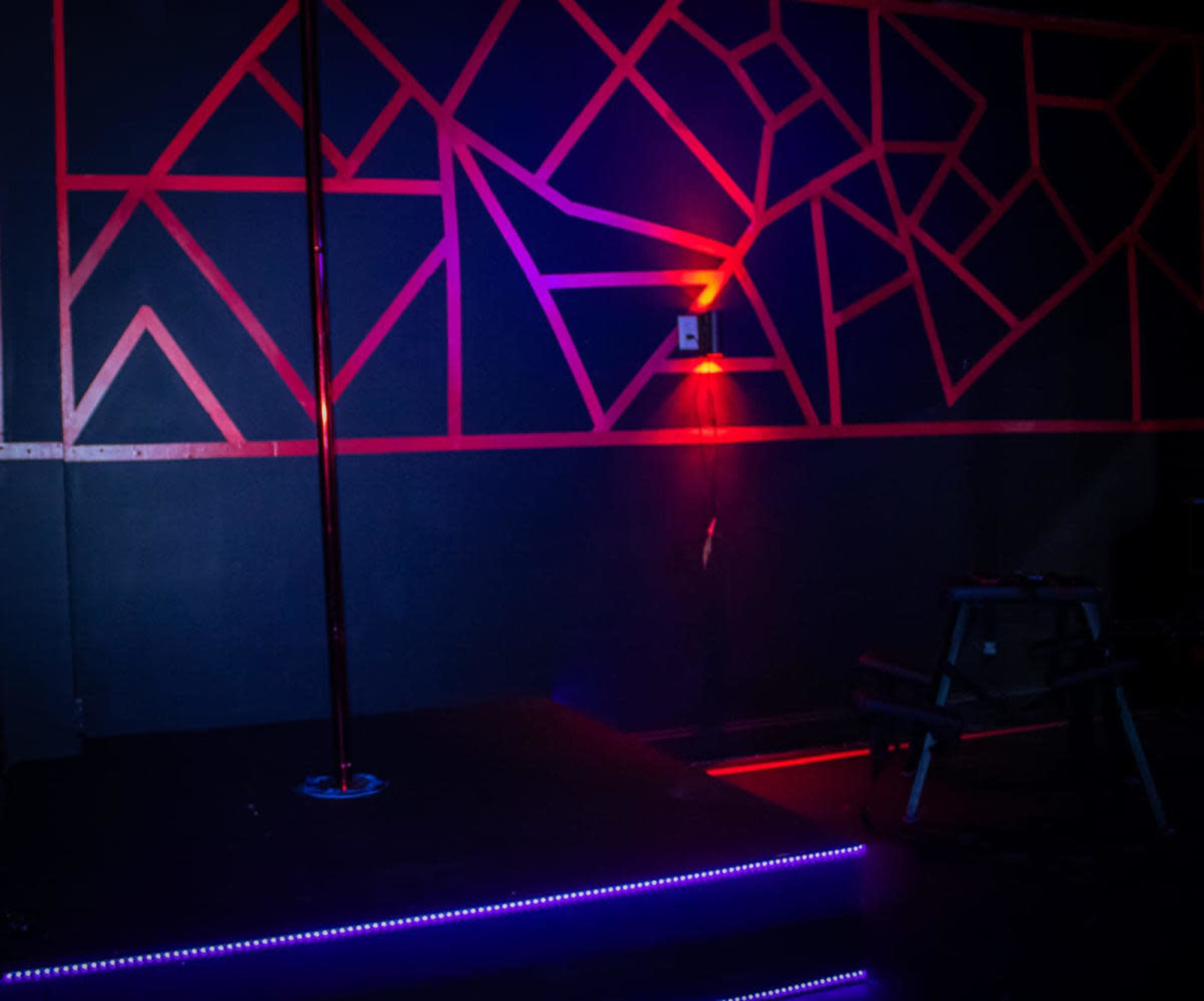 The image shows a dimly lit room with a centrally positioned pole, surrounded by a geometric-patterned wall illuminated by red and purple lights.