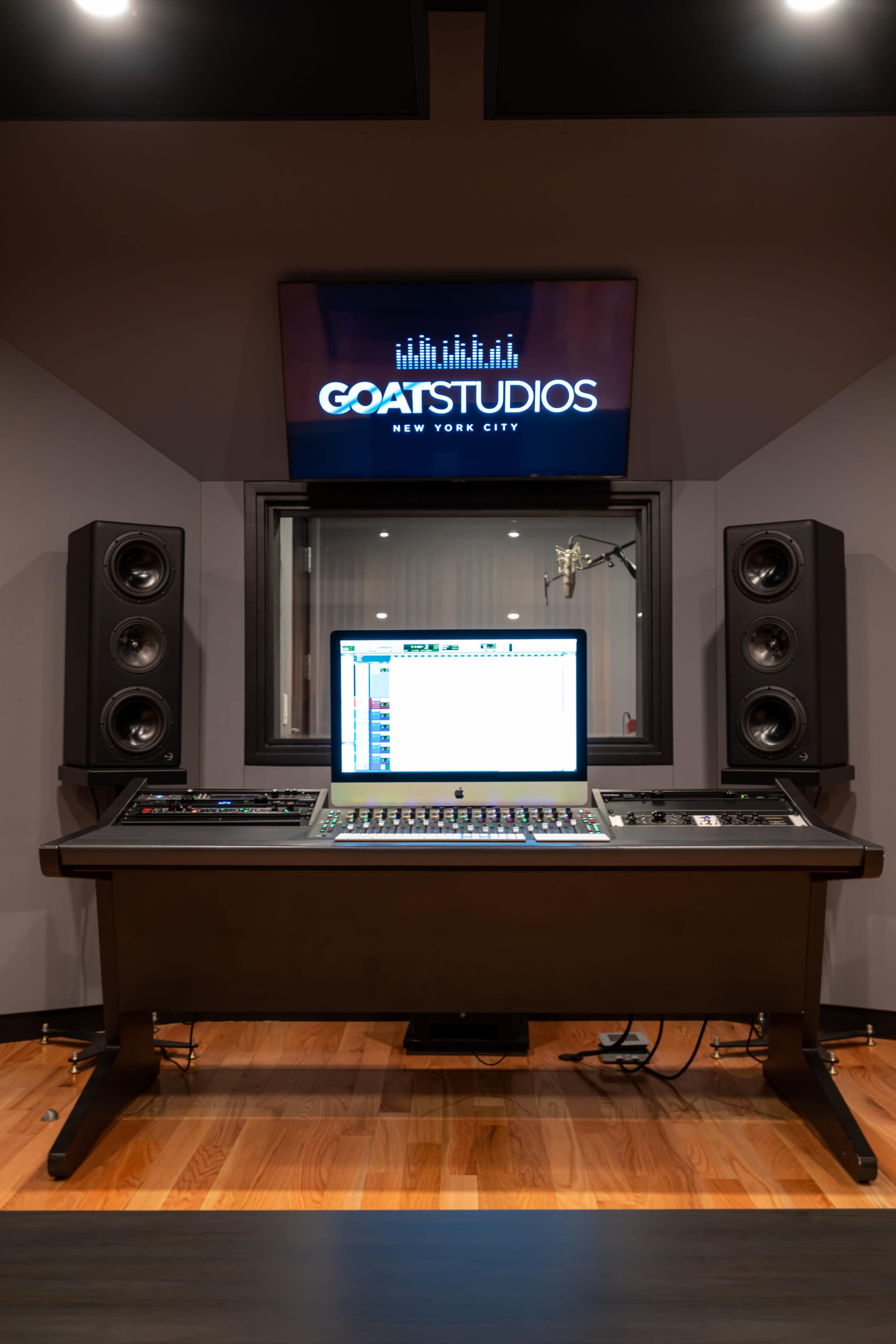 A recording studio with a large mixing console, a computer screen displaying audio software, and studio monitors on either side, all set against a backdrop featuring a logo for GOAT Studios.