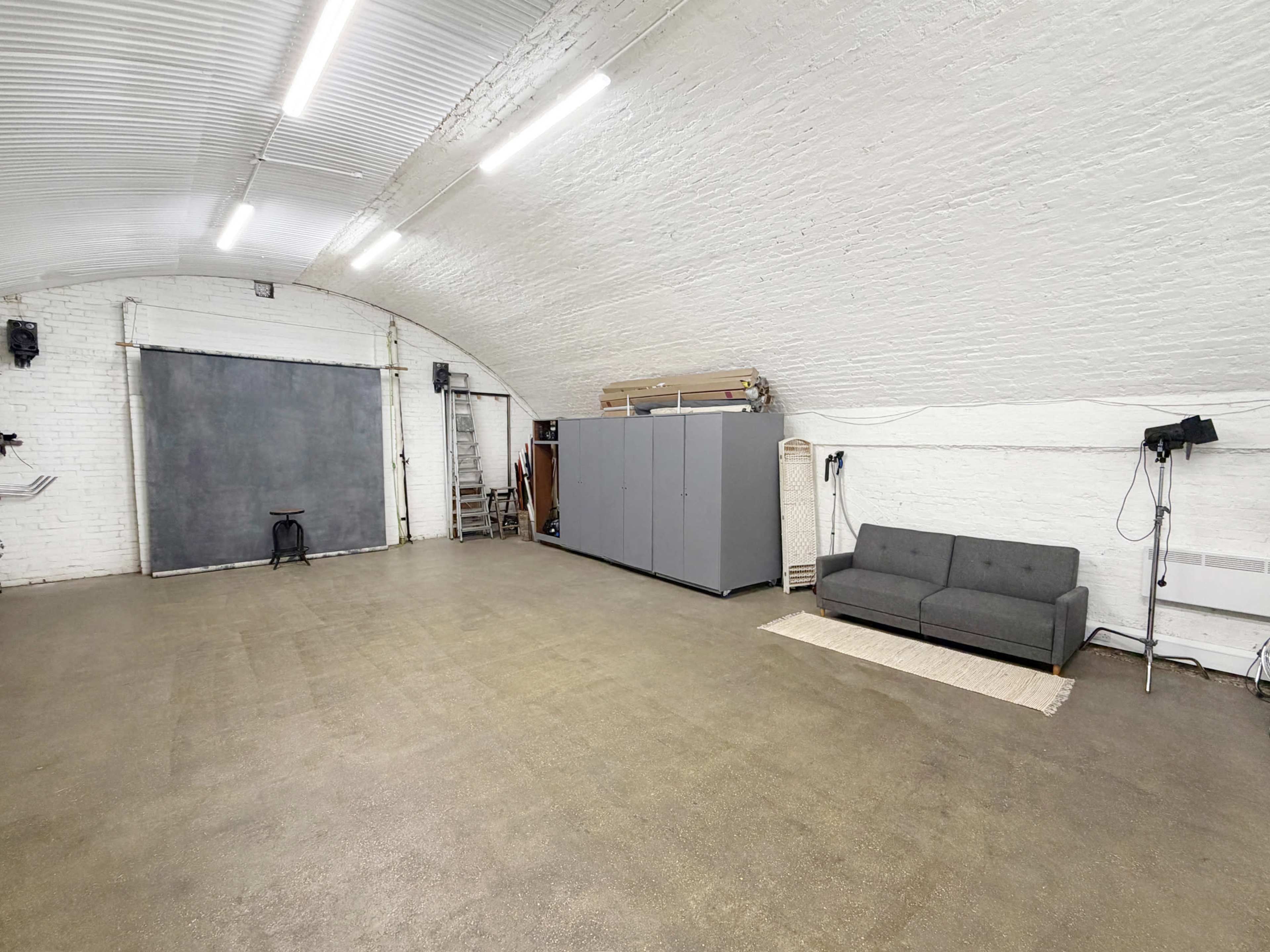 The image shows a spacious, empty room with white brick walls, a grey storage unit, a sofa, and a backdrop setup for photography.