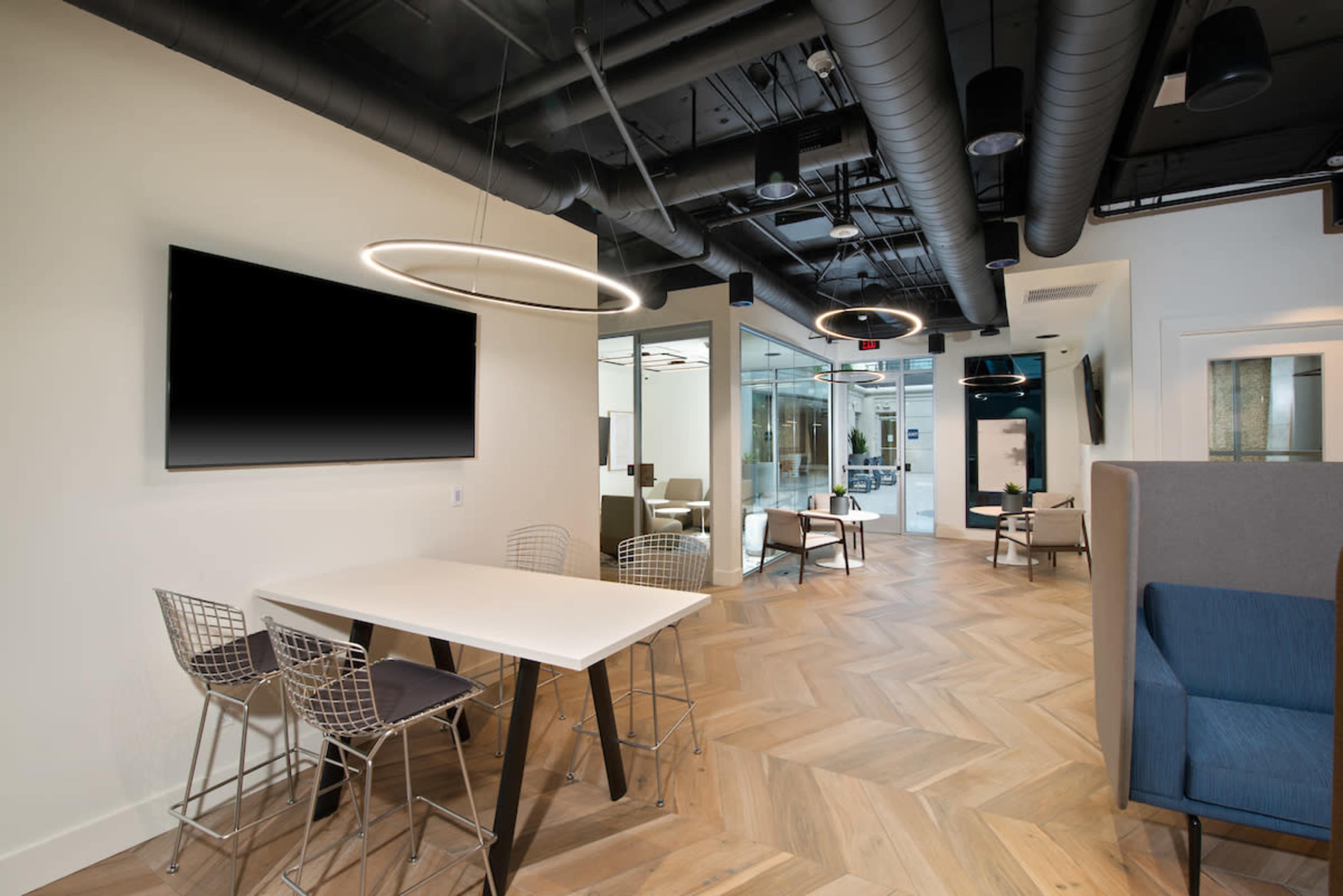 The image shows a modern office interior featuring a communal workspace with a table and chairs, sleek lighting, and an open layout with glass-walled conference rooms.