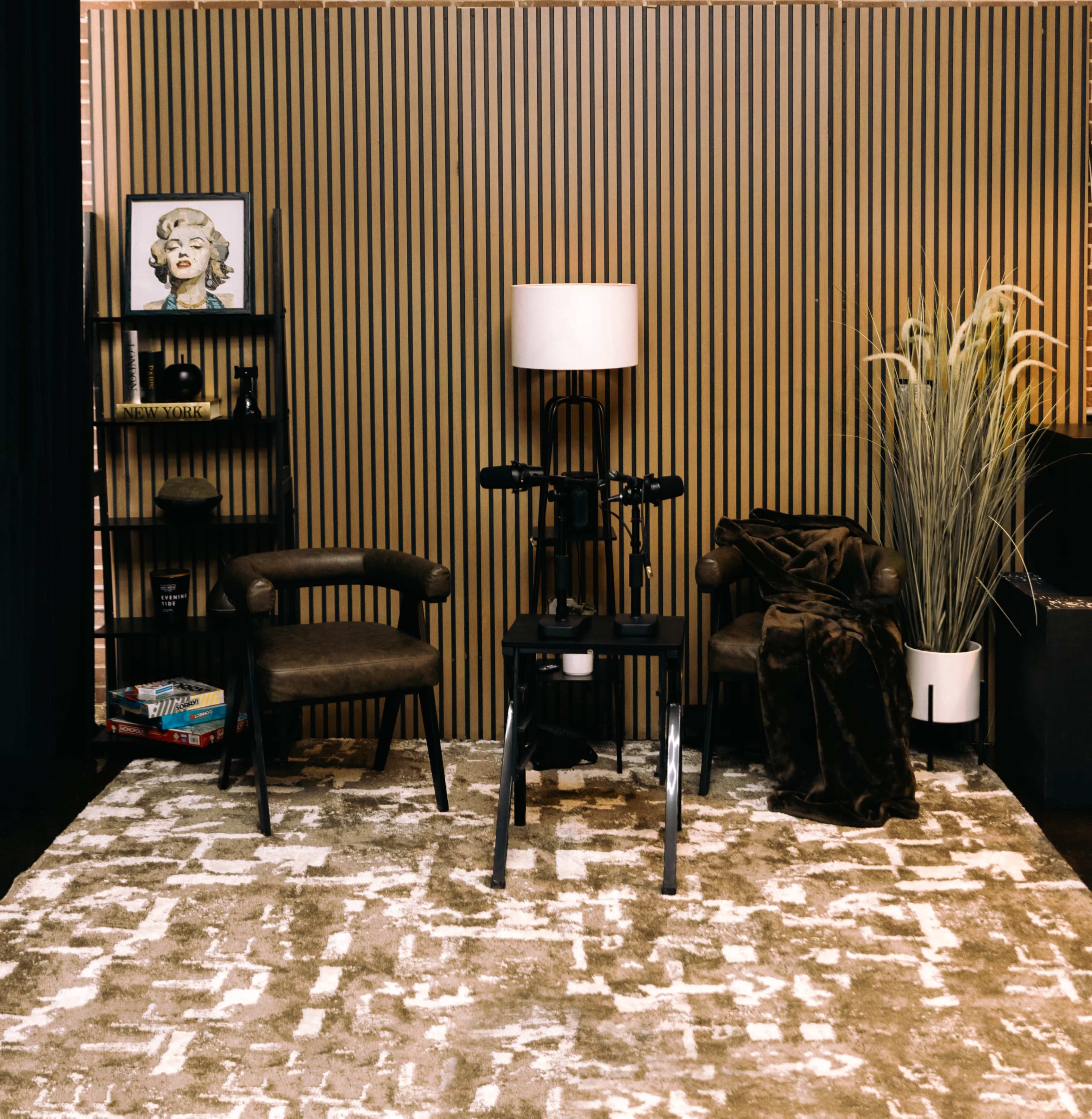 The scene features a cozy interior with two chairs facing a small table, a lamp, and a shelf against a striped wooden wall, complemented by a rug and decorative plants.