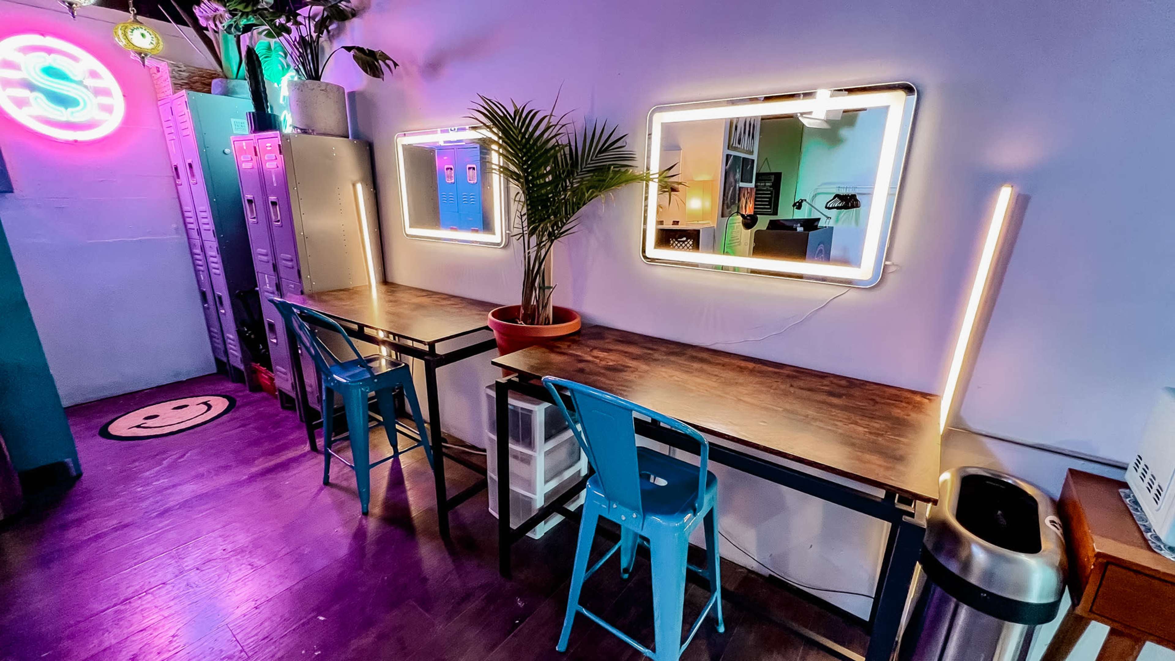 The image shows a cozy workspace with two metal tables and blue chairs, illuminated by neon lights and featuring potted plants against a backdrop of lockers.