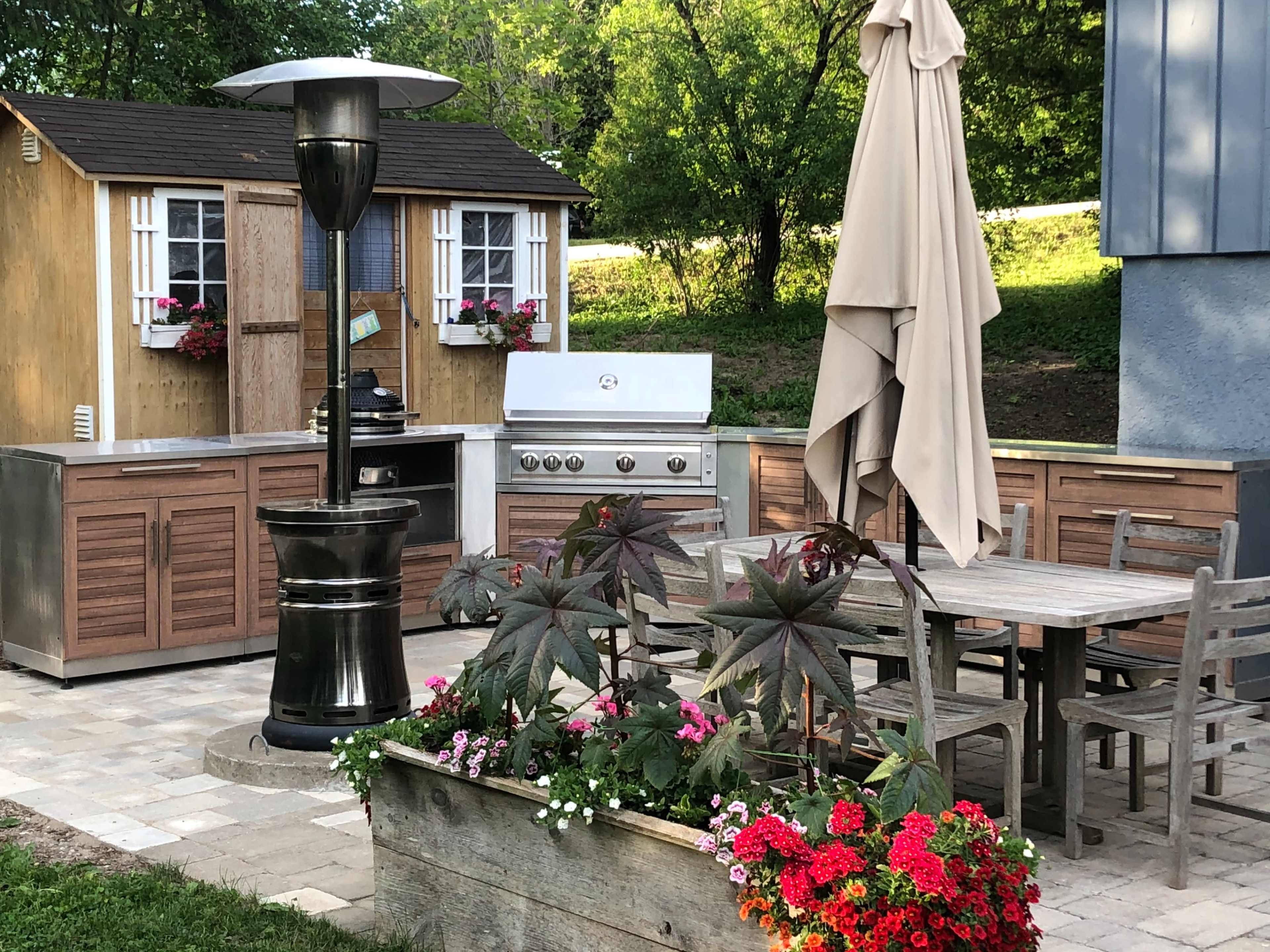 An outdoor kitchen area with a stainless steel grill, wooden cabinets, a patio table with chairs, a large umbrella, and a decorative planter filled with flowers.