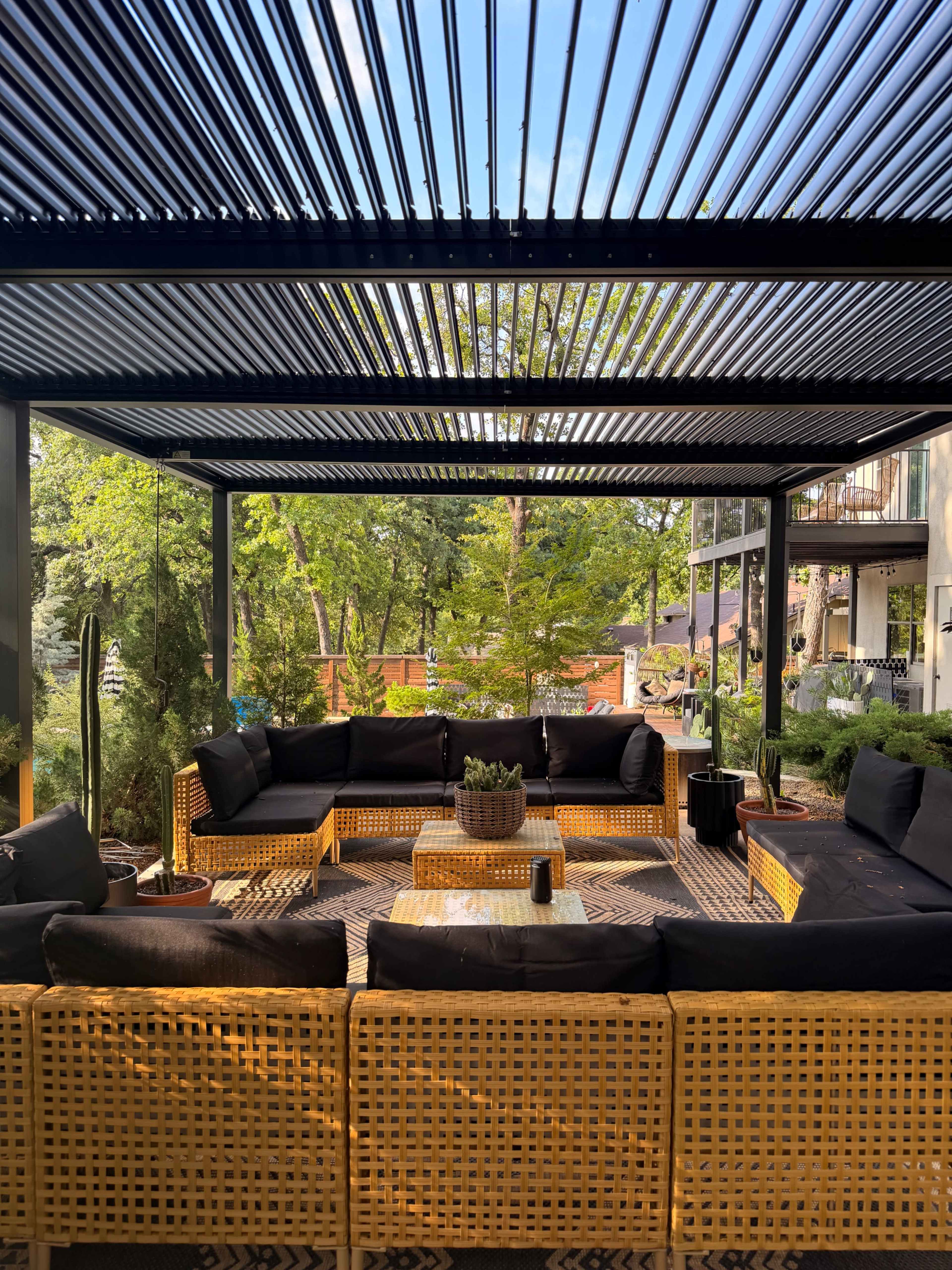 The image shows a shaded outdoor seating area with a large, arranged sectional sofa, a coffee table, and green plants surrounding it.
