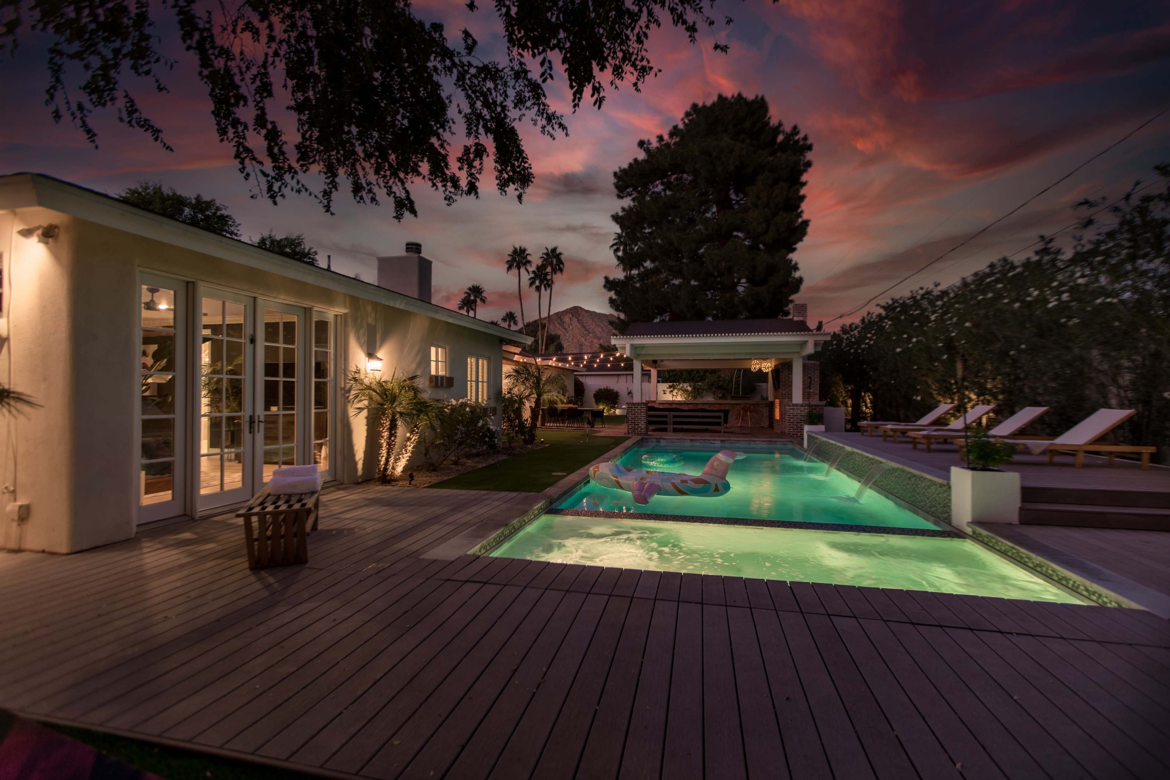 A modern backyard with a swimming pool, wooden decking, lounge chairs, and a sunset sky in the background.