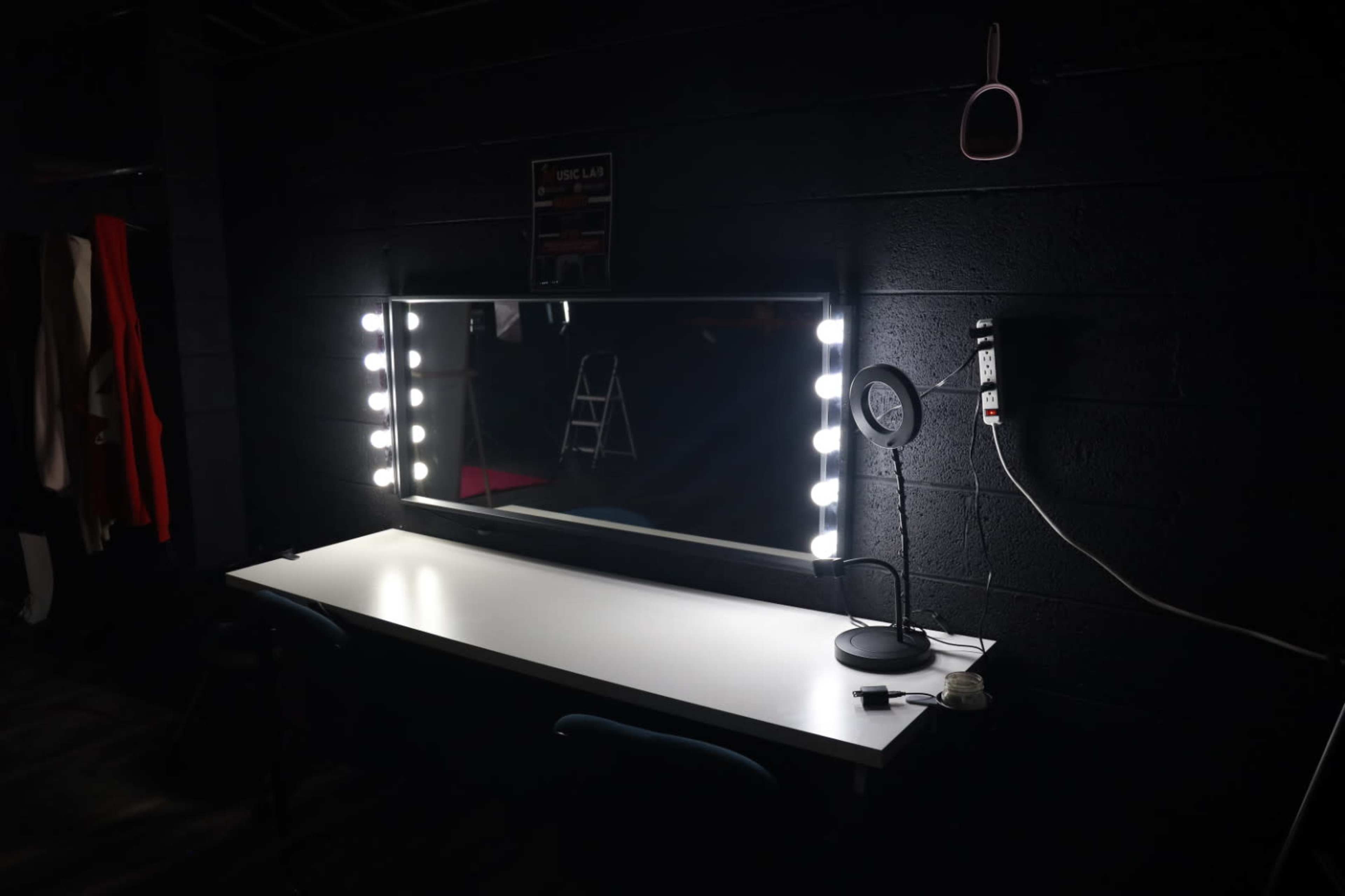A dimly lit room featuring a large mirror with surrounding lights and a desk with a lamp and a chair.