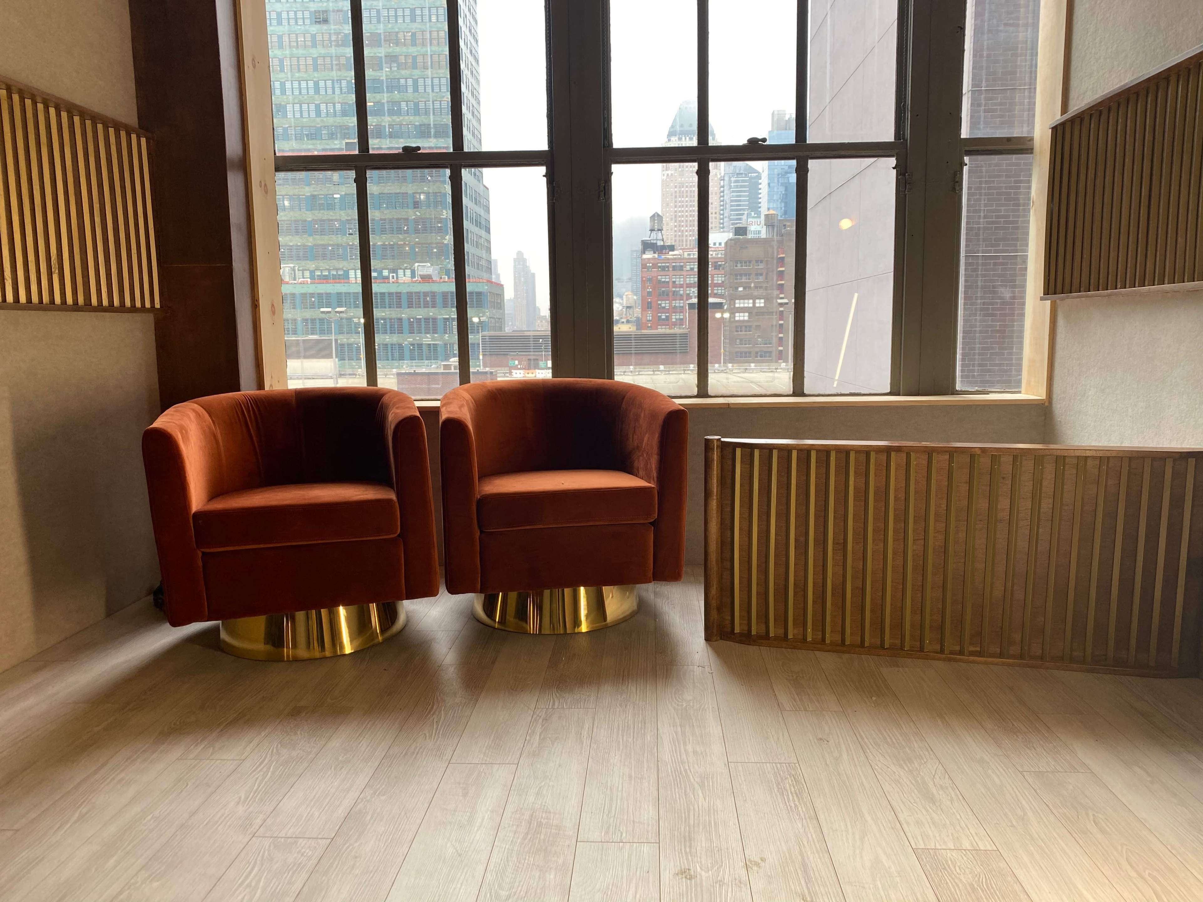 Two orange velvet chairs with gold bases are positioned near a large window overlooking a city skyline.