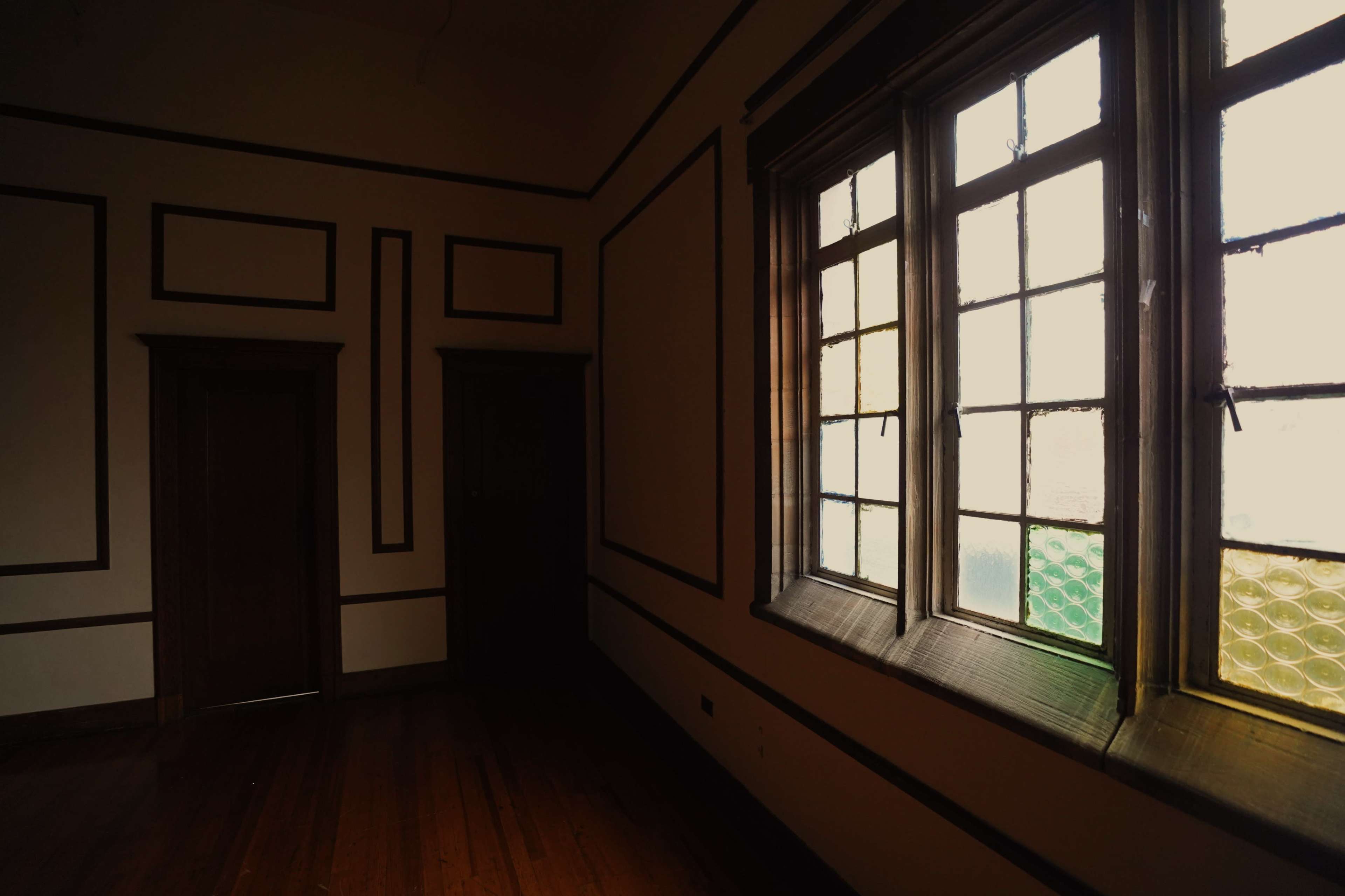 The image shows a dimly lit room with wooden floors, featuring a large window with multiple panes and decorative wall trim.