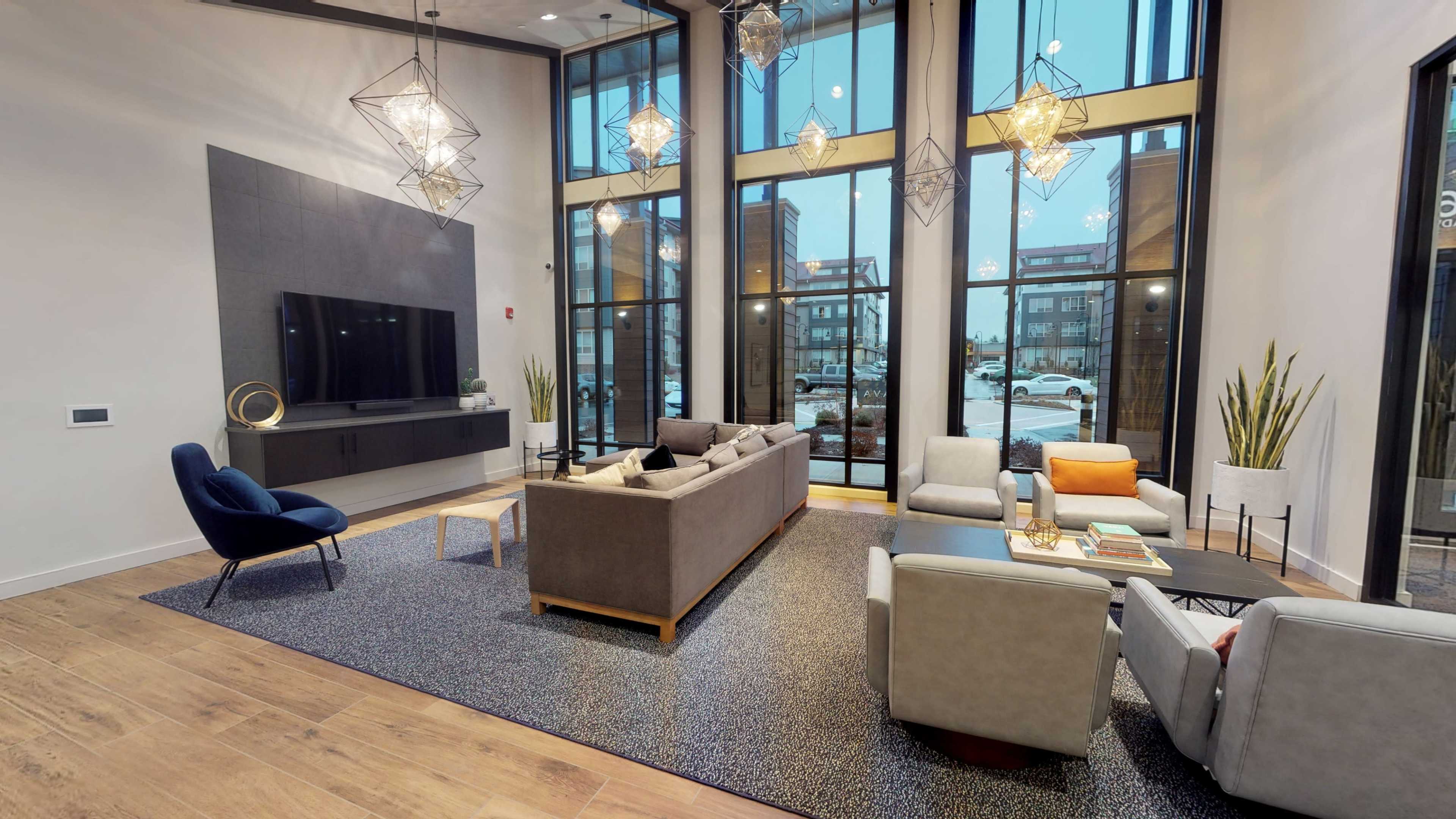 A modern lounge area featuring large windows, a television mounted on a wall, stylish light fixtures, and a mix of seating arrangements with a coffee table.