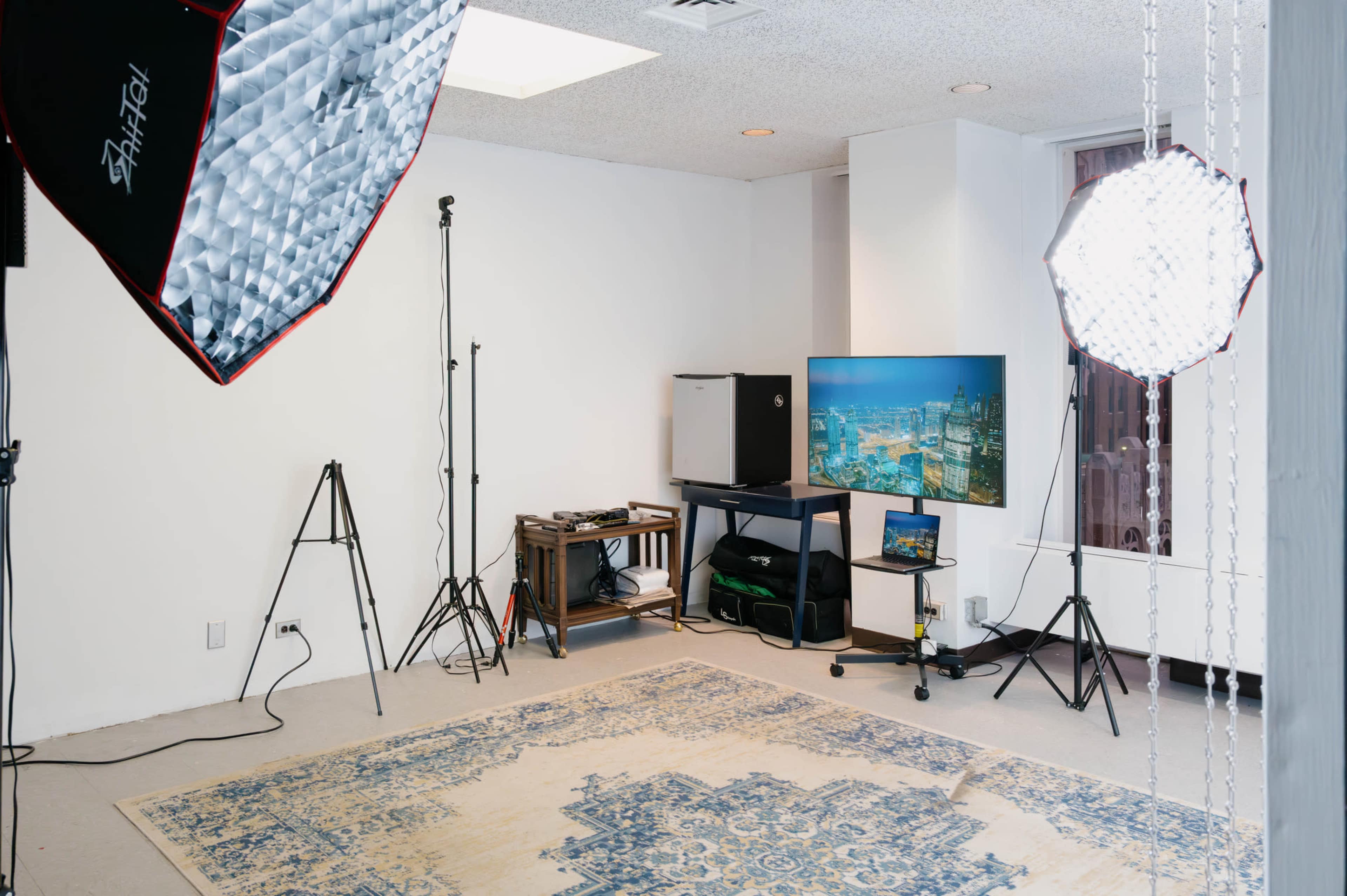 The image shows a studio space with a patterned rug, a television on a stand, and lighting equipment set up on tripods.