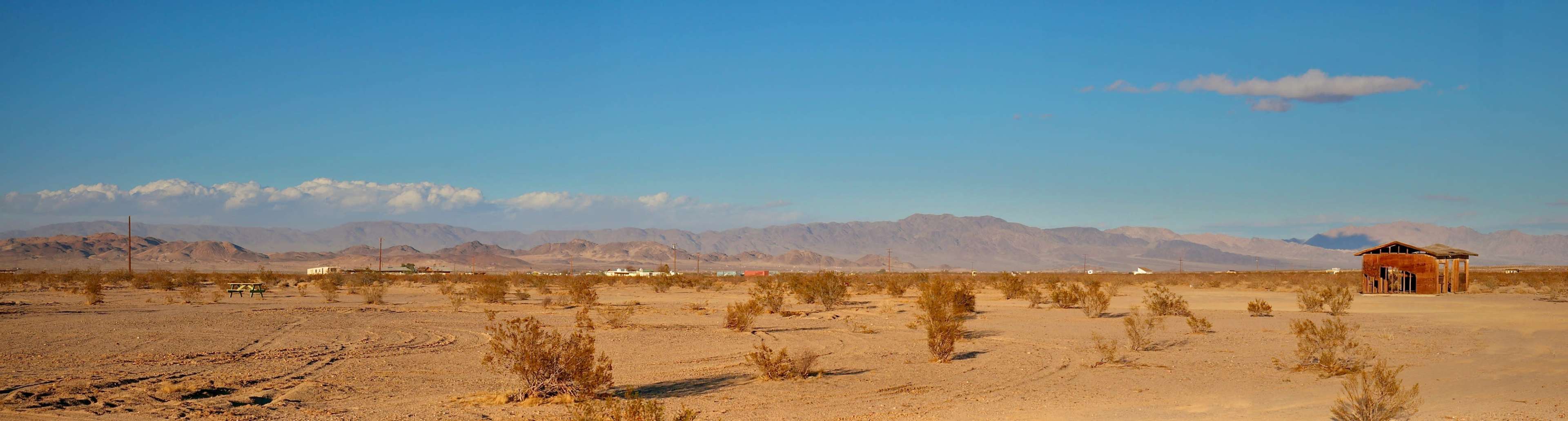 A sparse desert landscape features a small wooden structure against a backdrop of distant mountains and a clear blue sky.