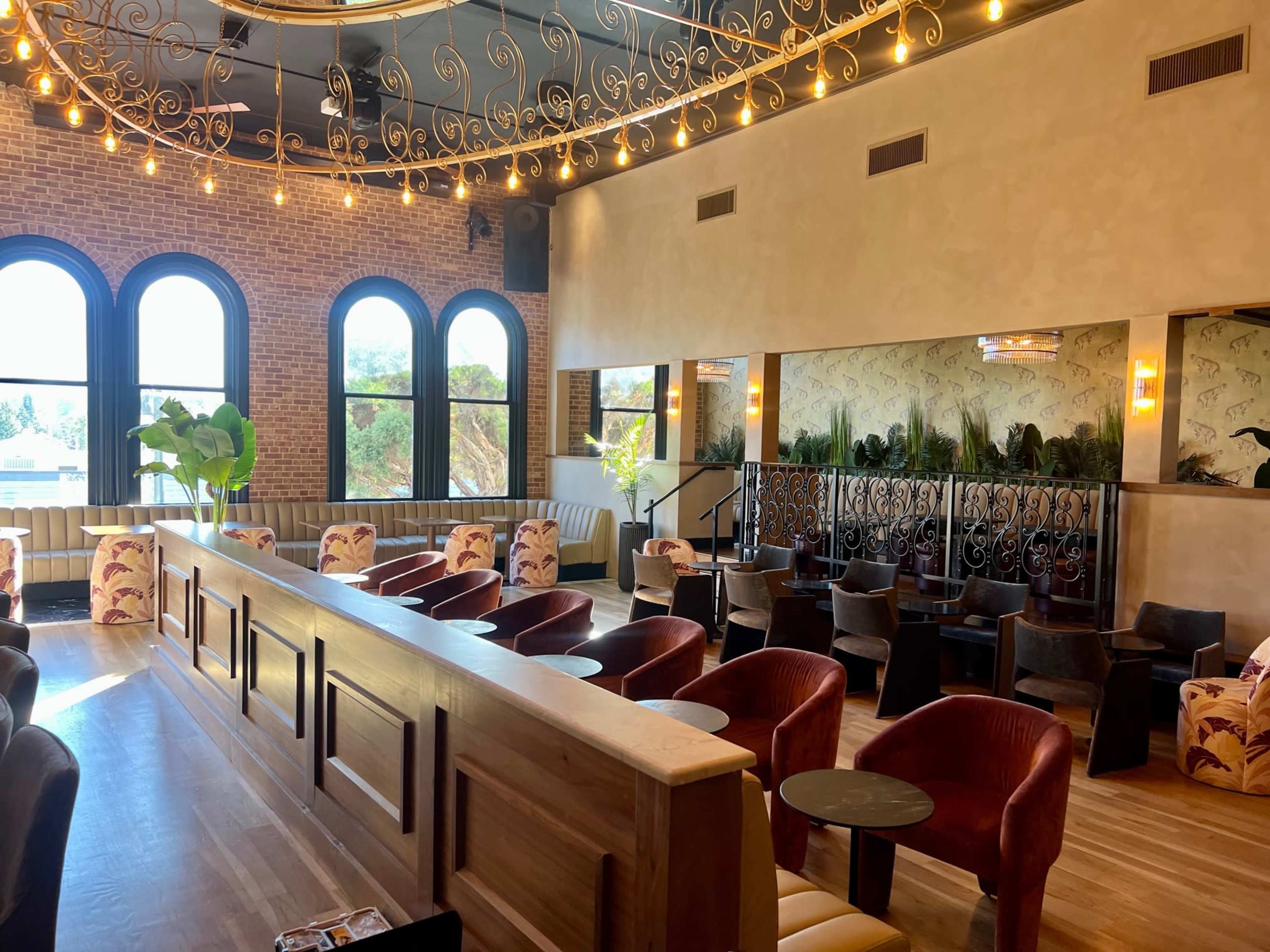 The image shows a stylish interior of a restaurant or lounge featuring wooden seating, large windows, and decorative lighting.