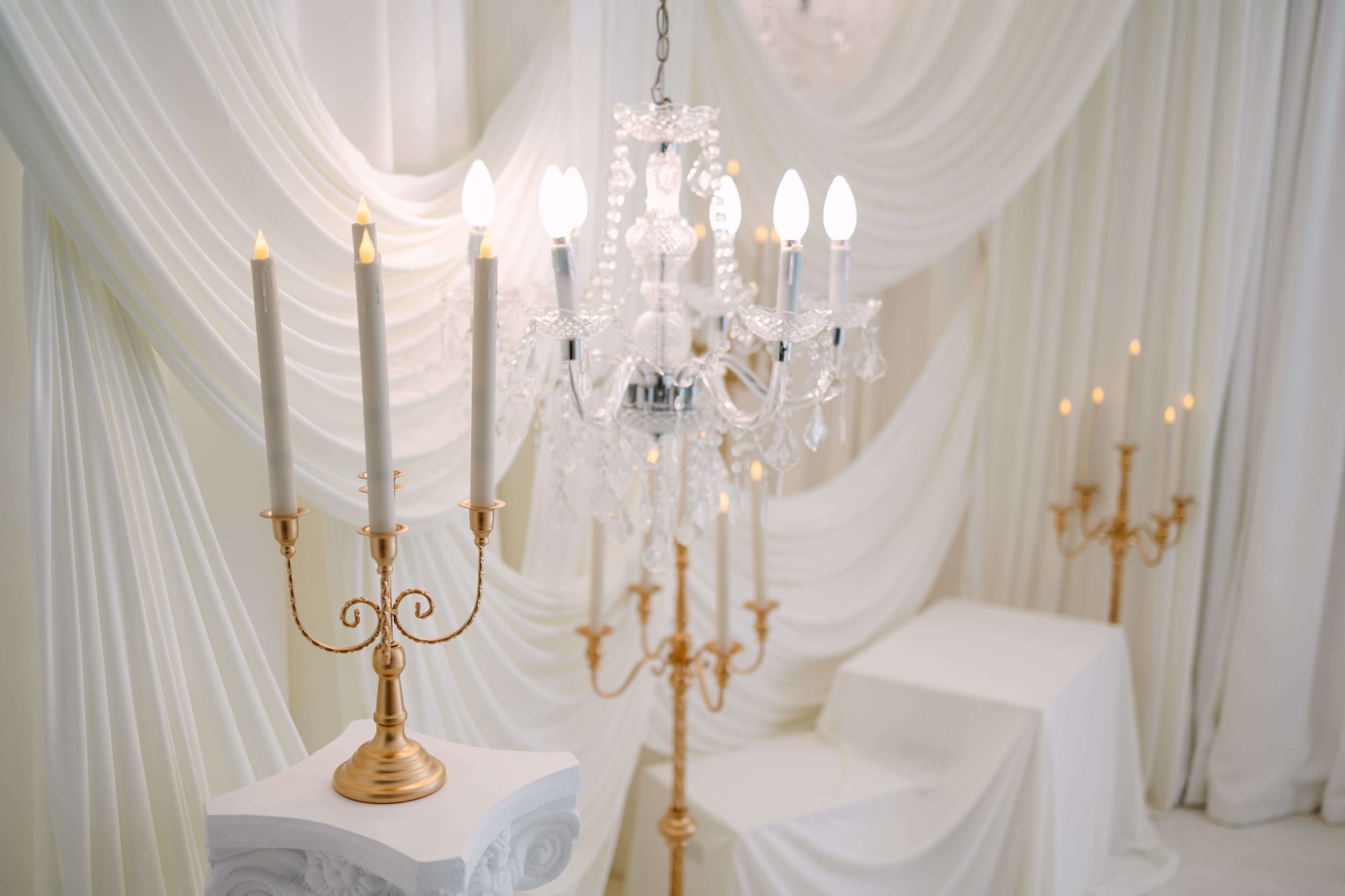 The image features an elegant chandelier hanging above a set of white draped fabric, accompanied by golden candle holders on pedestals.