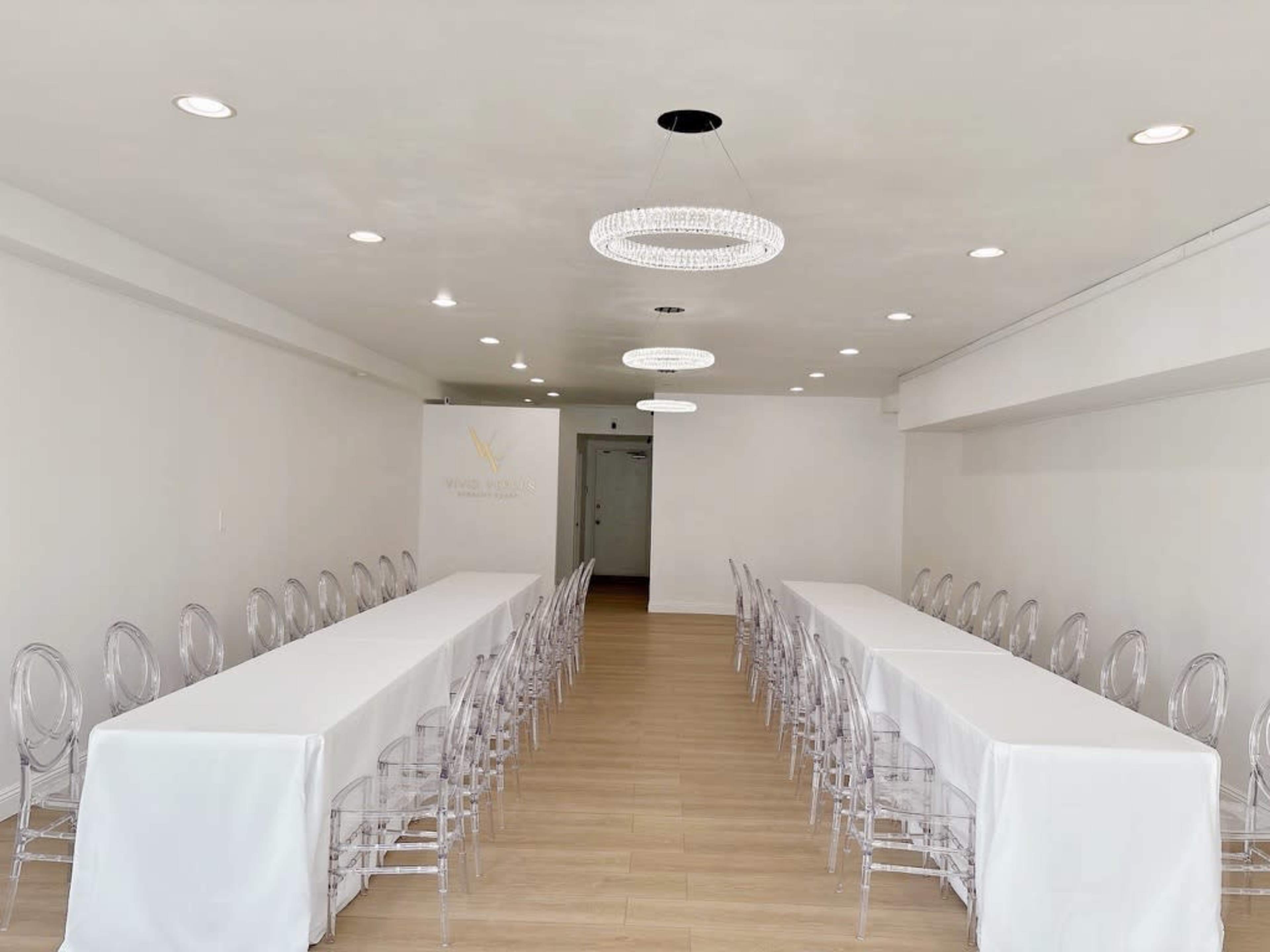 The image shows a bright, minimalist event space with two long tables covered in white tablecloths and surrounded by clear acrylic chairs.