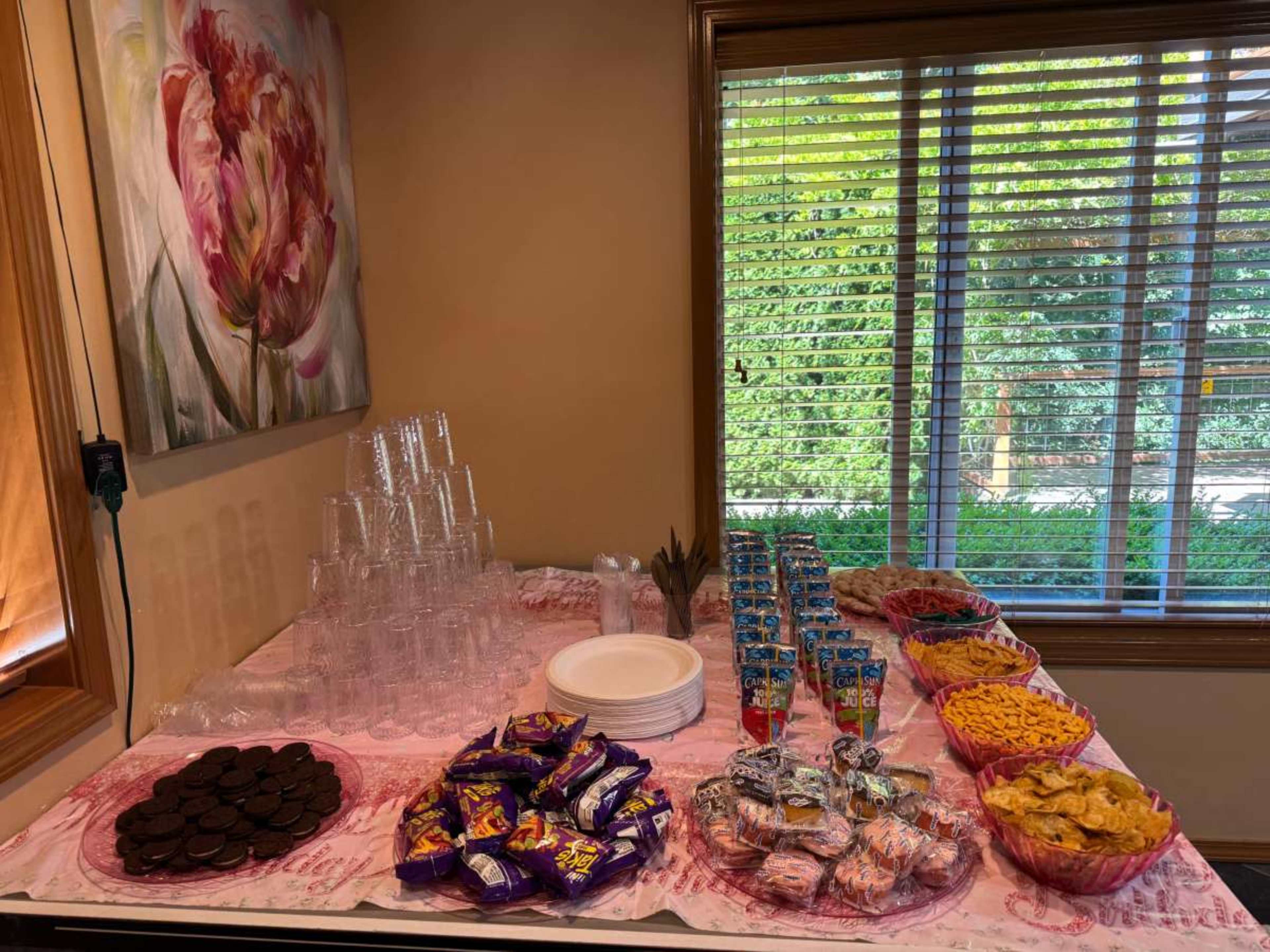 A table is displayed with an assortment of snacks, drinks, and plastic cups arranged neatly in a room with a large window.