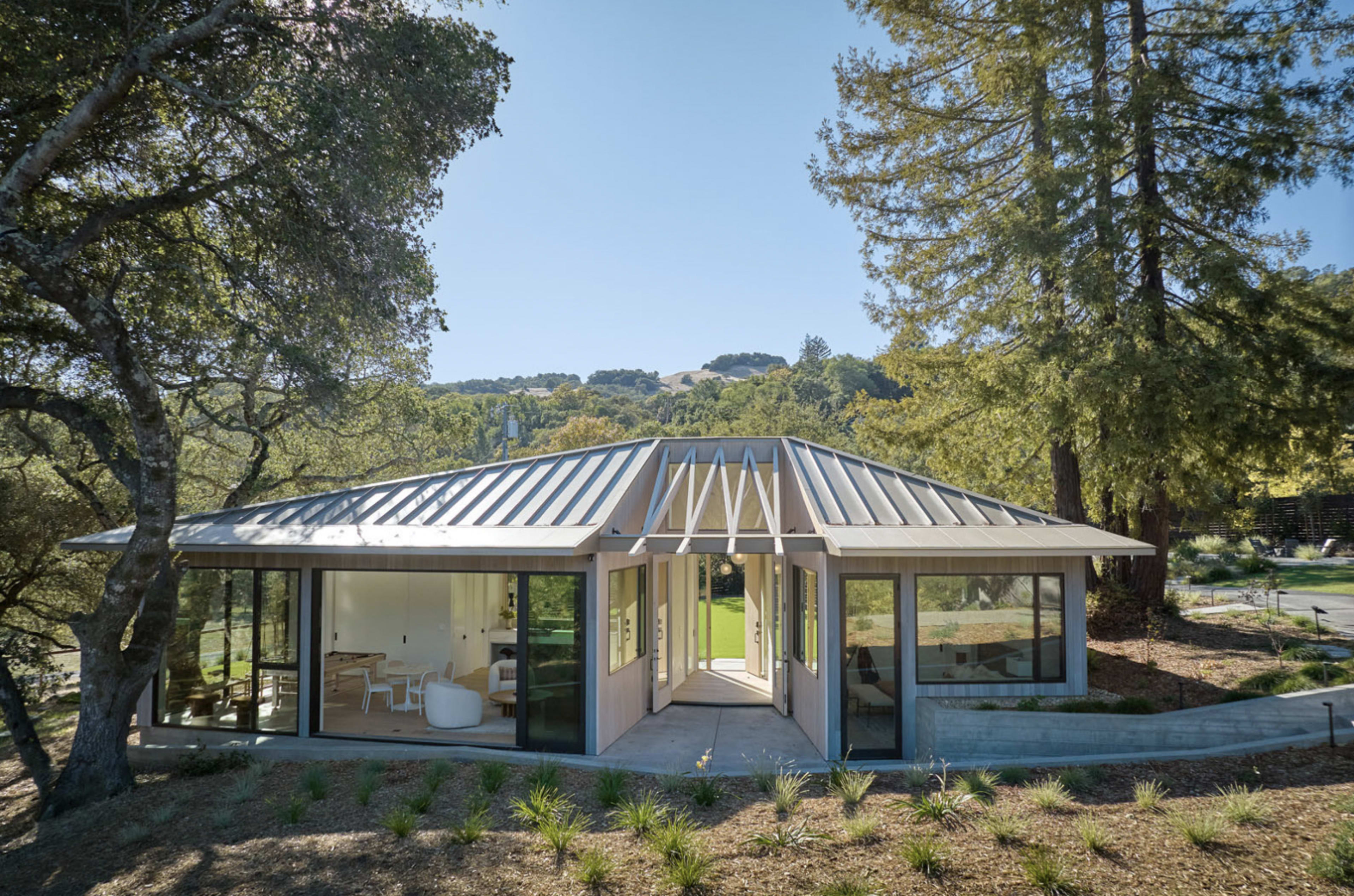A modern house with large glass windows and a corrugated metal roof is surrounded by trees and greenery.