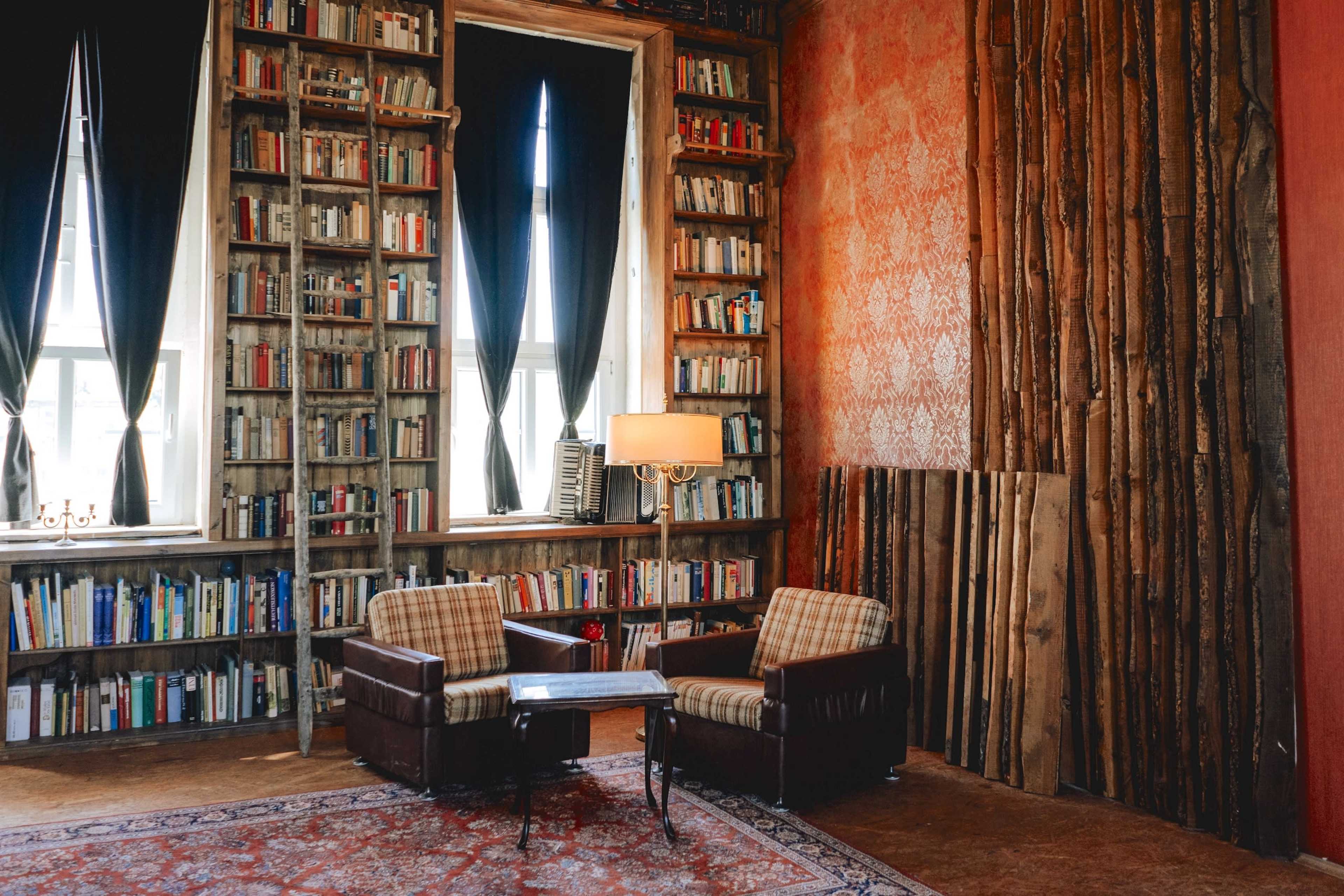 A cozy reading nook features two armchairs, a small table, and bookshelves filled with books, set against a backdrop of textured walls and large windows with dark curtains.