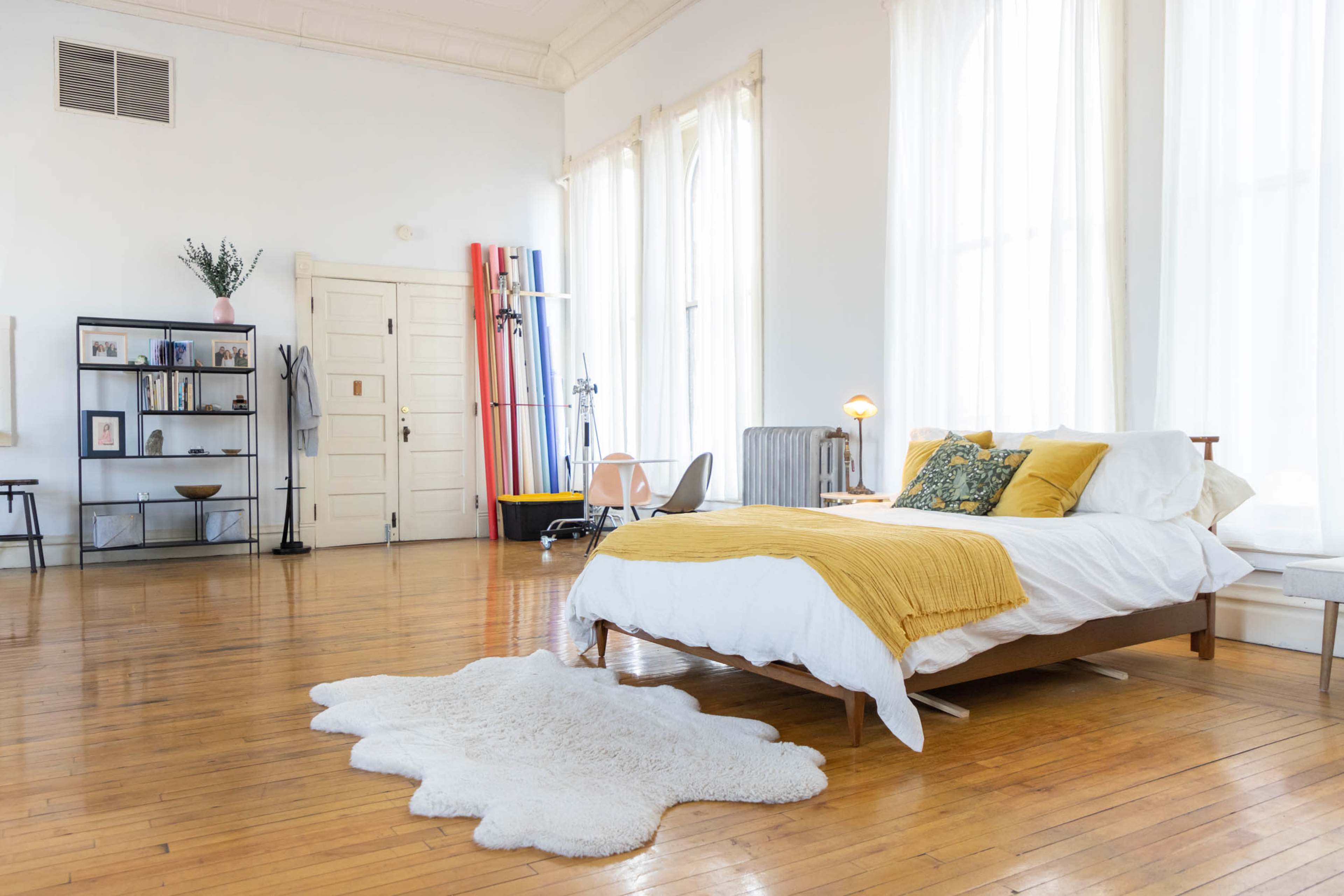 The image shows a bright, minimalistic bedroom with a wooden bed, a fluffy white rug on the polished wooden floor, and large windows allowing natural light to fill the space.
