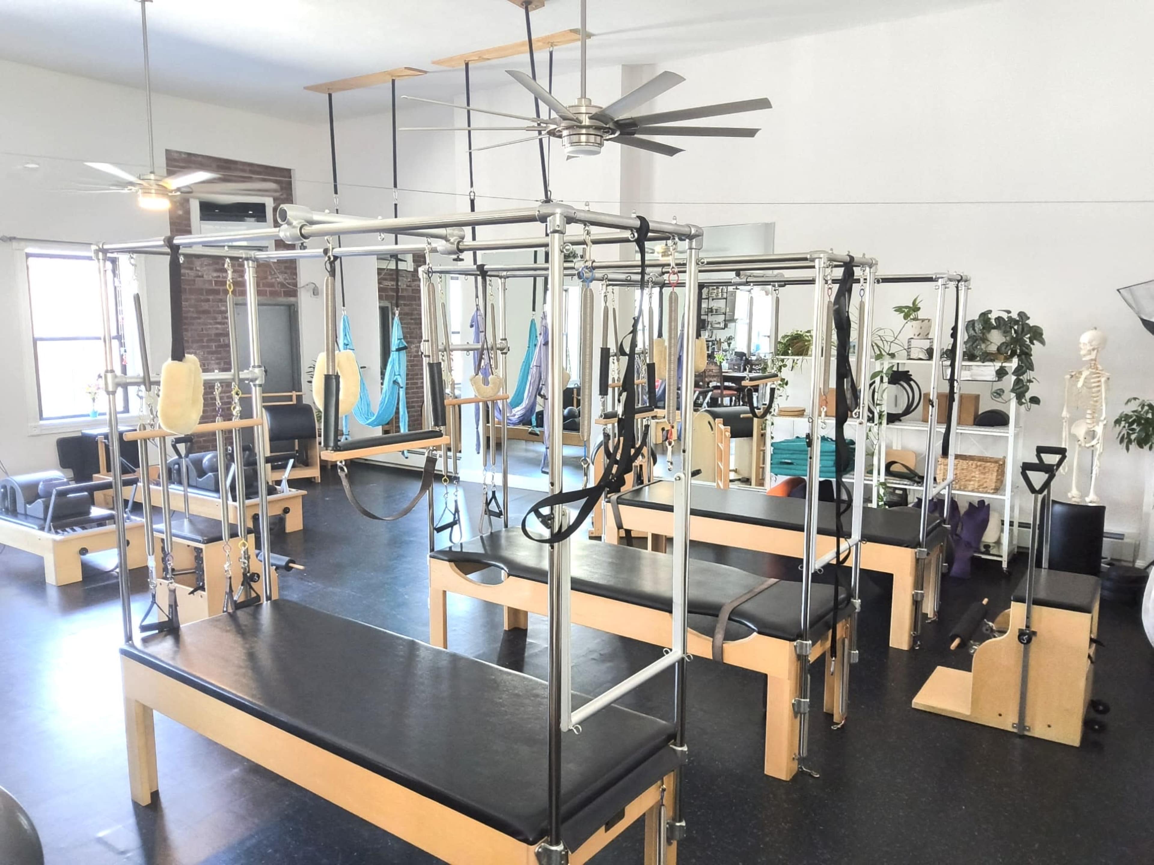 The image shows a Pilates studio filled with several reformer machines arranged in a spacious room with large windows and ceiling fans.