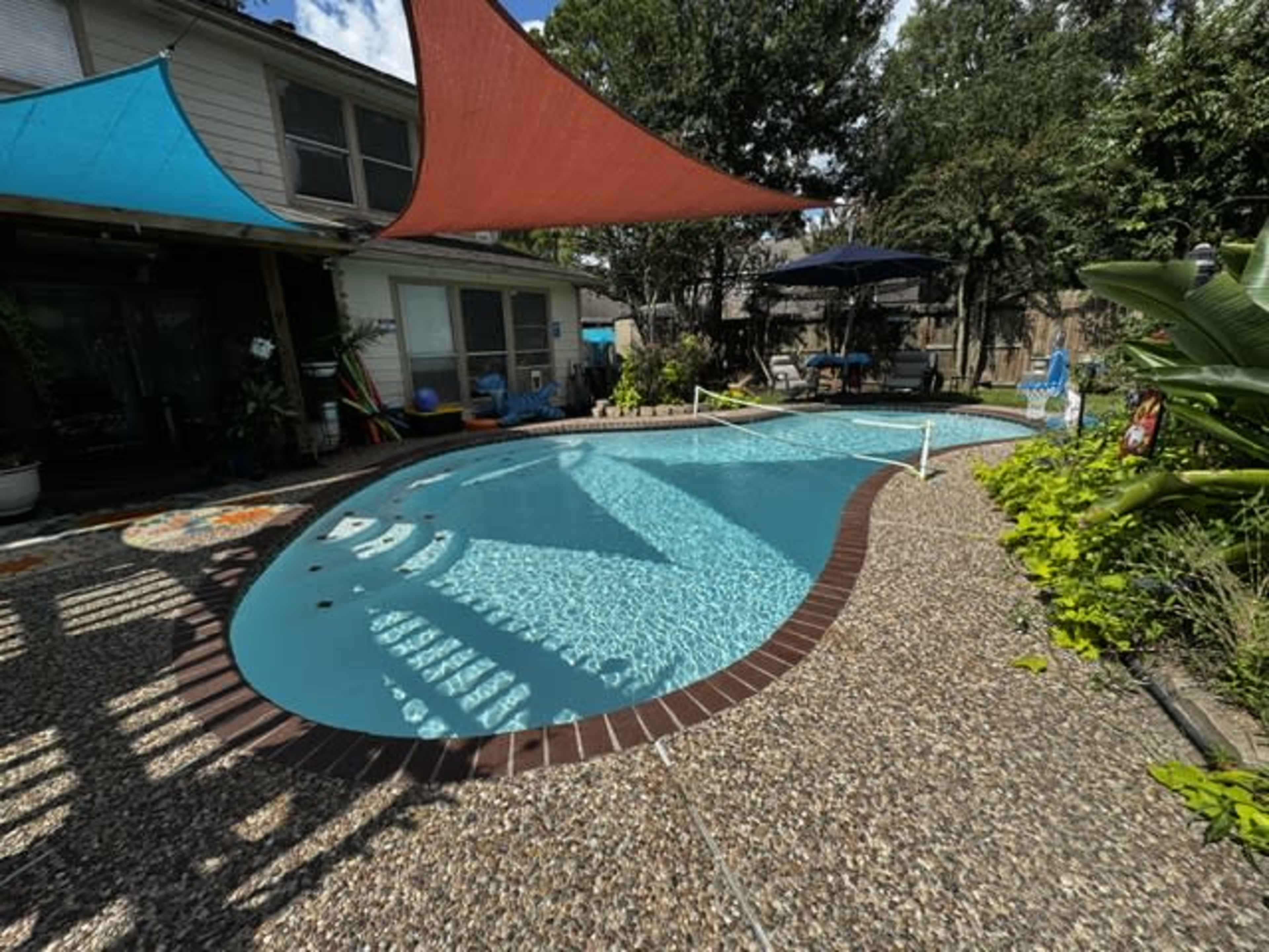 The image shows a backyard pool with decorative shade sails, surrounded by a landscaped area featuring tropical plants and outdoor seating.