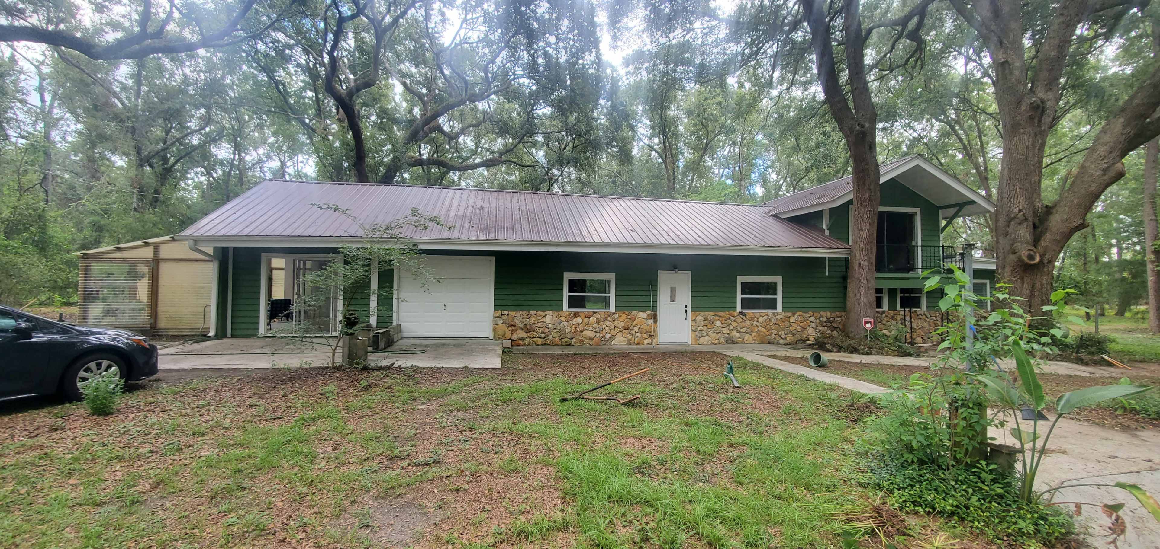 A green two-story house with a stone facade and a metal roof is surrounded by trees and a grassy area.