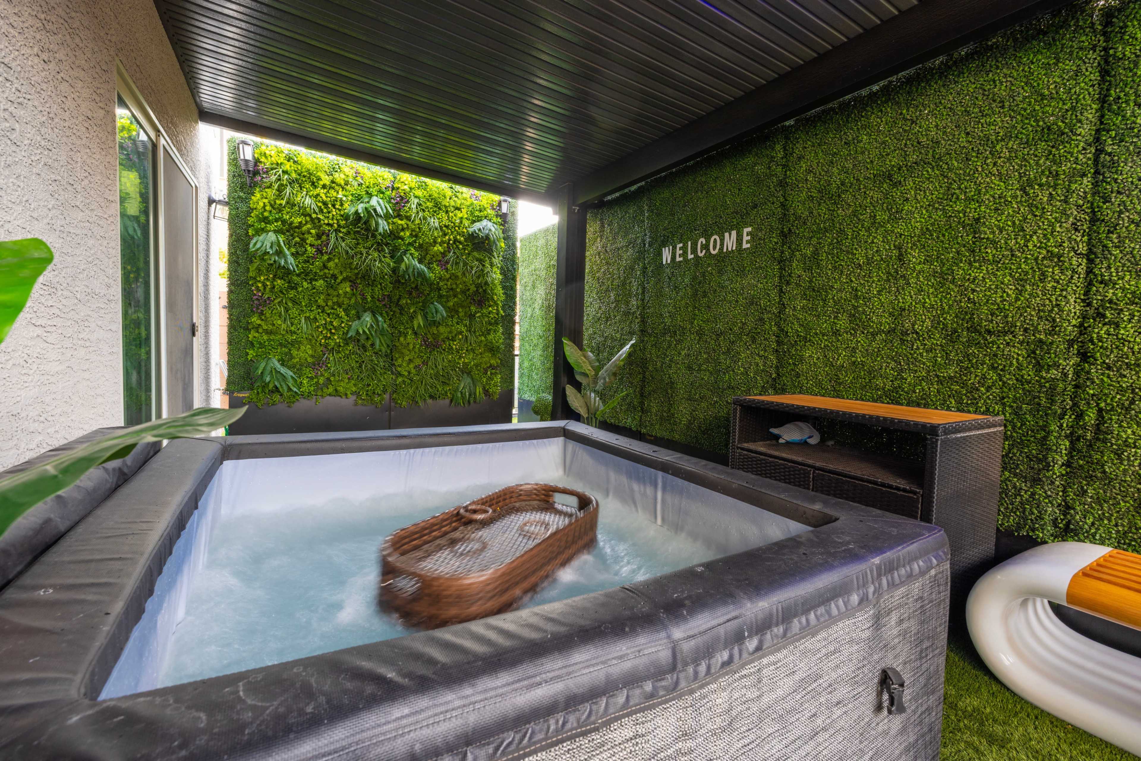 The image shows a hot tub with bubbling water surrounded by a green wall of plants and a welcome sign.
