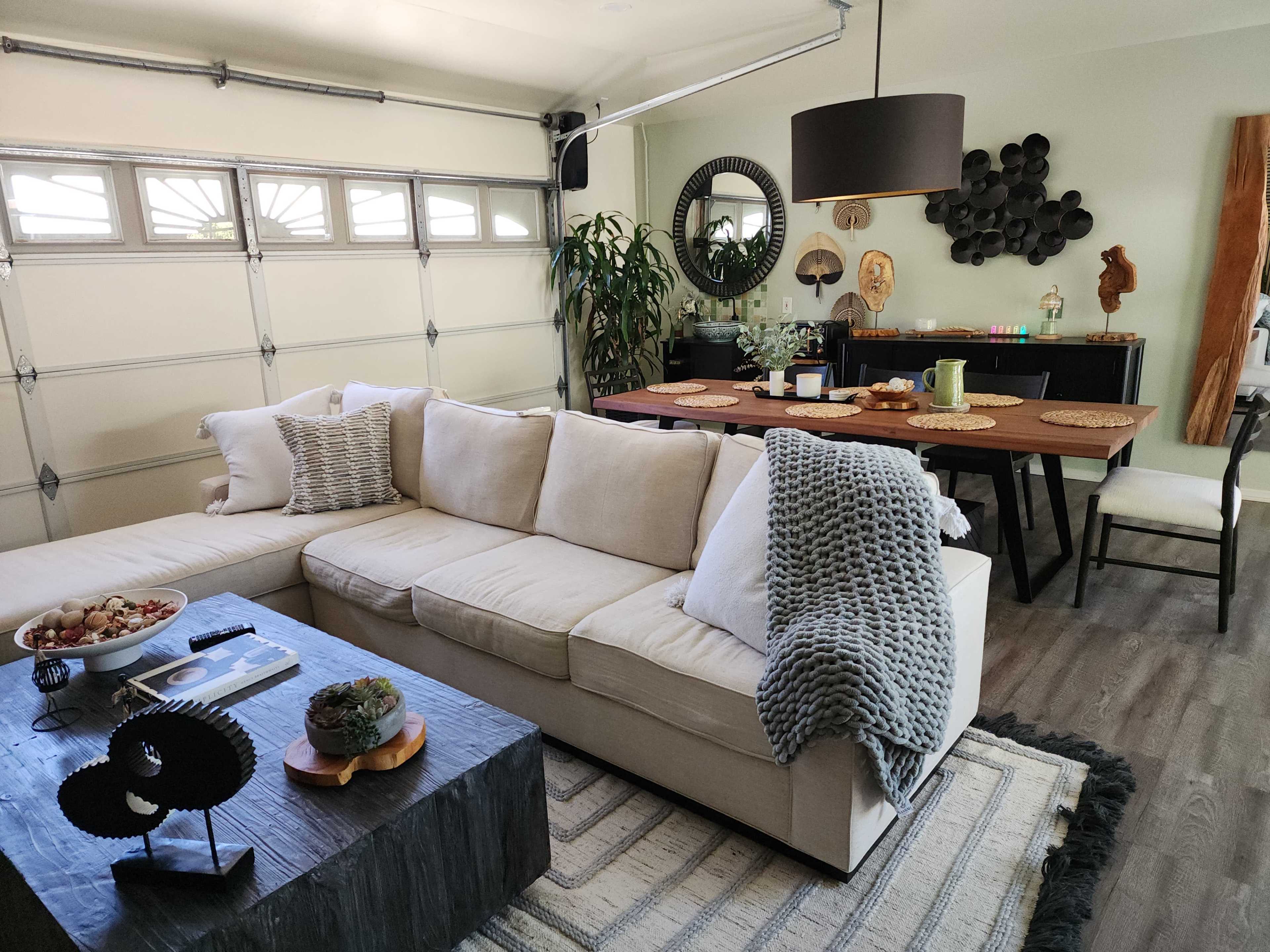 A spacious living area features a large L-shaped beige sofa with a gray throw blanket, a dark wooden coffee table, and a dining table set for several people.