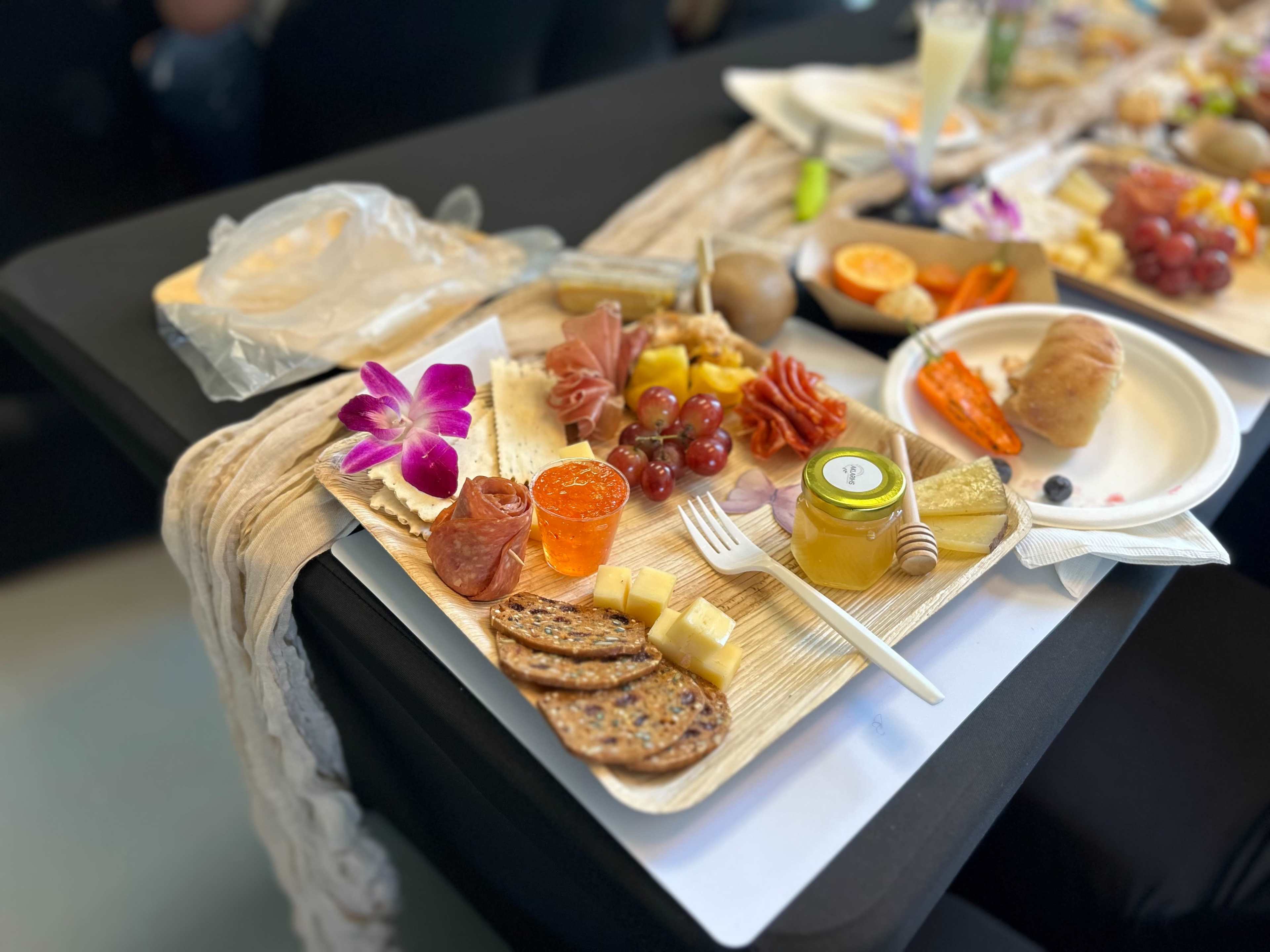 A wooden tray is filled with an assortment of cheeses, cured meats, fruits, and garnishes, accompanied by a small container of honey and a decorative flower.
