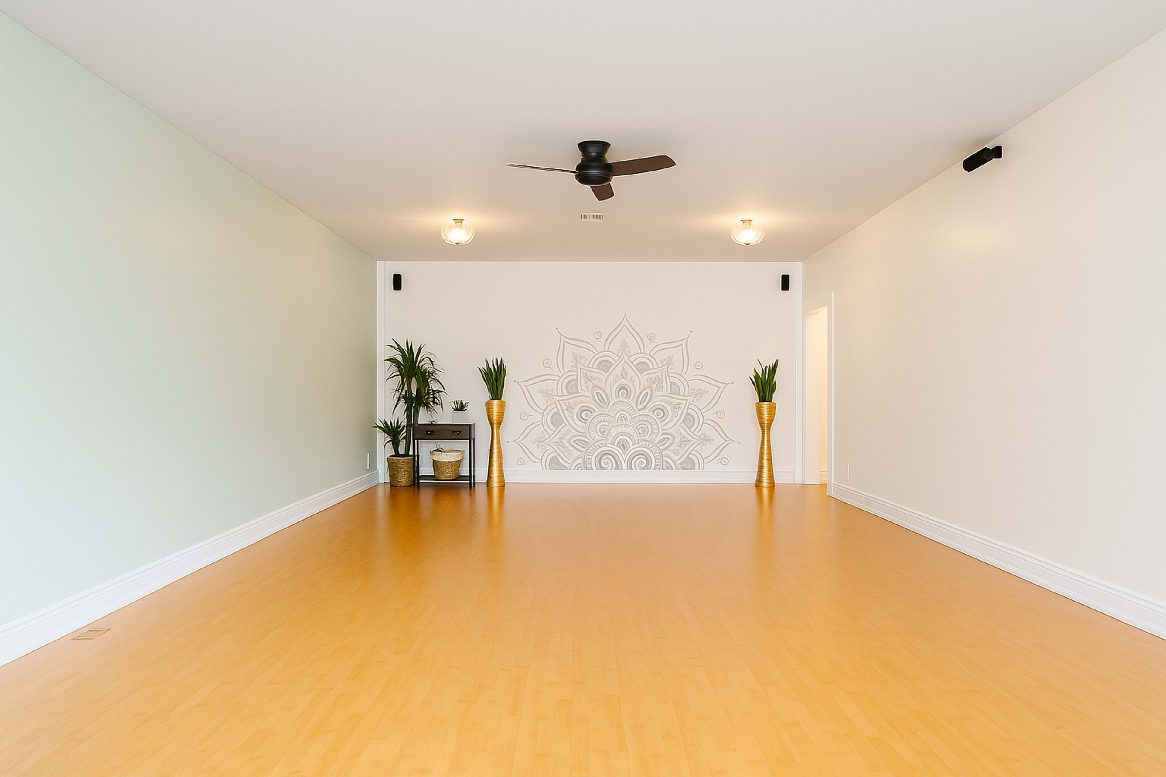 The image shows a spacious, well-lit room with bamboo flooring, featuring a decorative wall mural, two potted plants, and a ceiling fan.