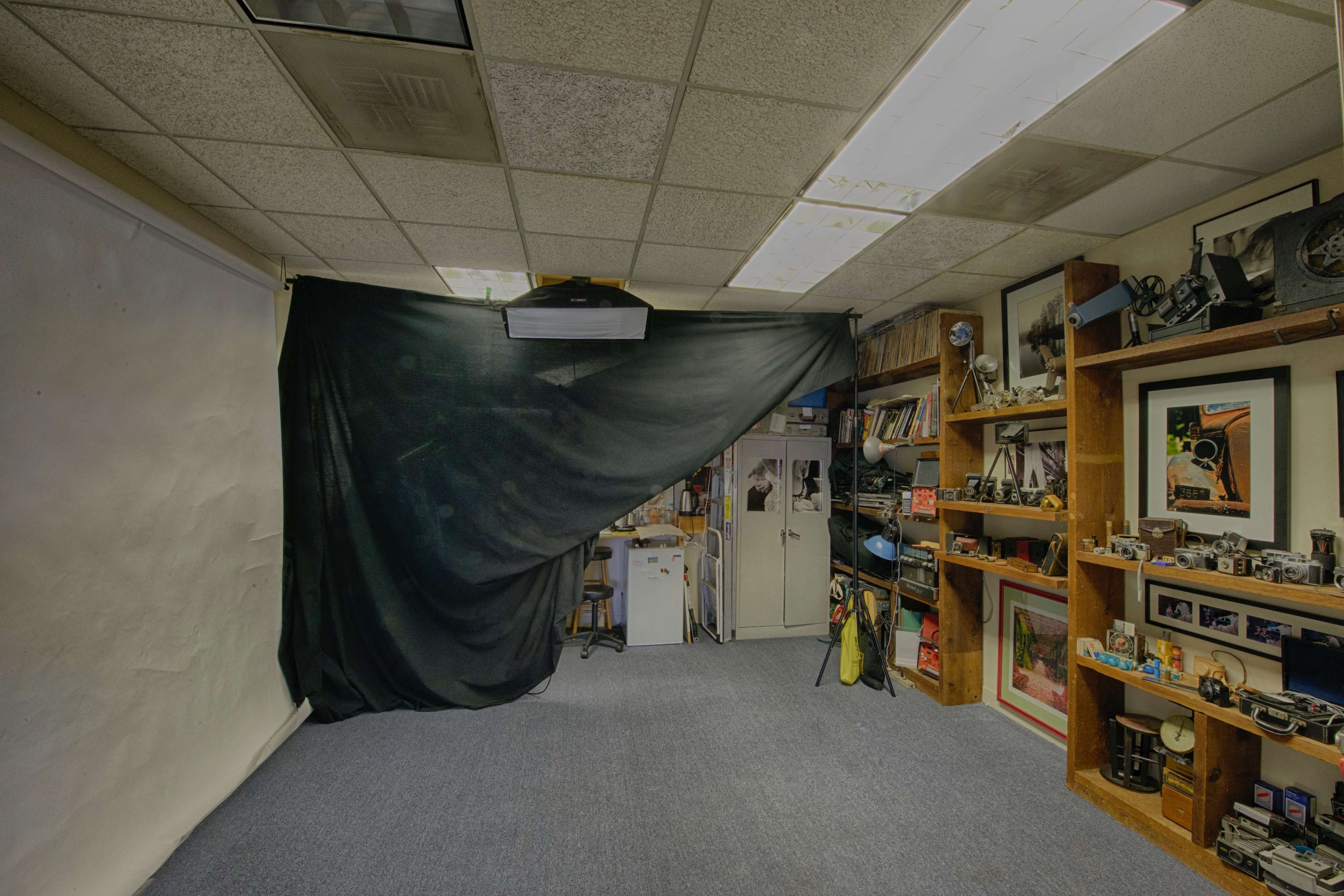 The image shows a photography studio with a black curtain draped across a wall, shelves filled with cameras, and a projector mounted on the ceiling.