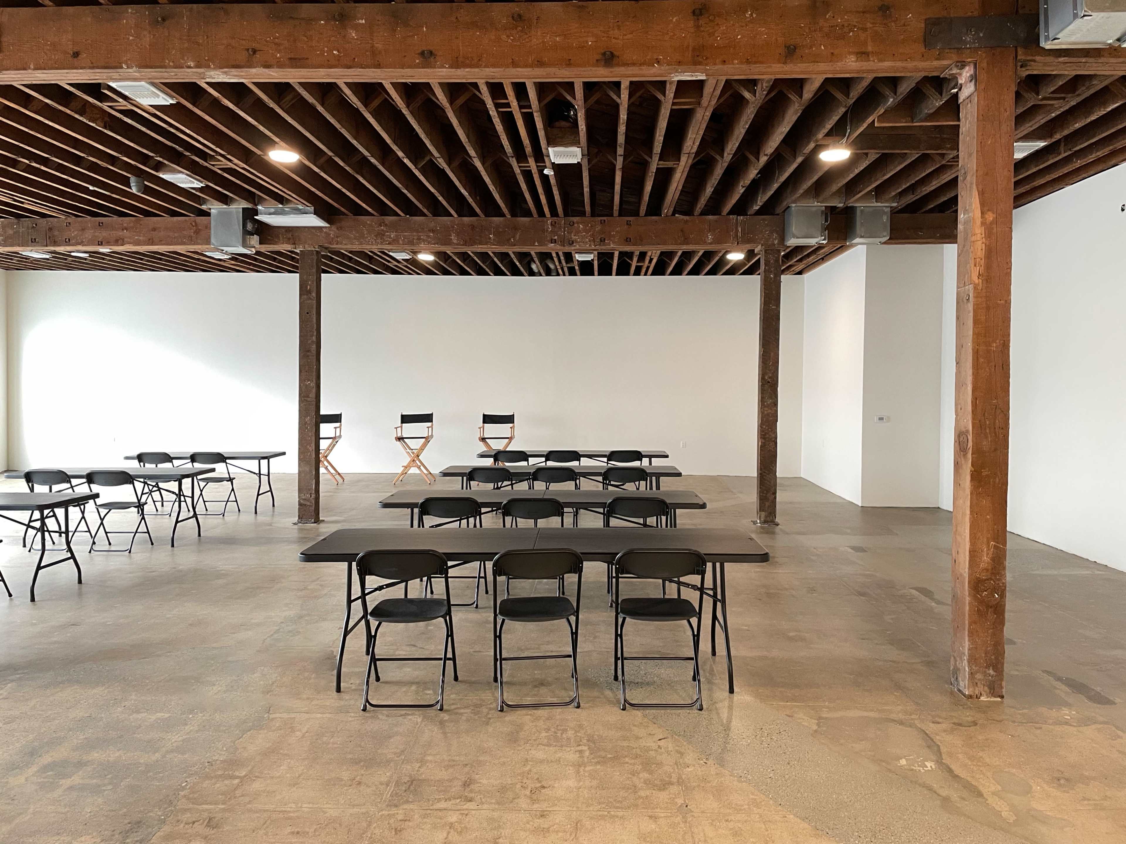 A spacious room with wooden beams overhead, featuring several black tables and chairs arranged in a minimalist layout, with three director's chairs positioned against a white wall.