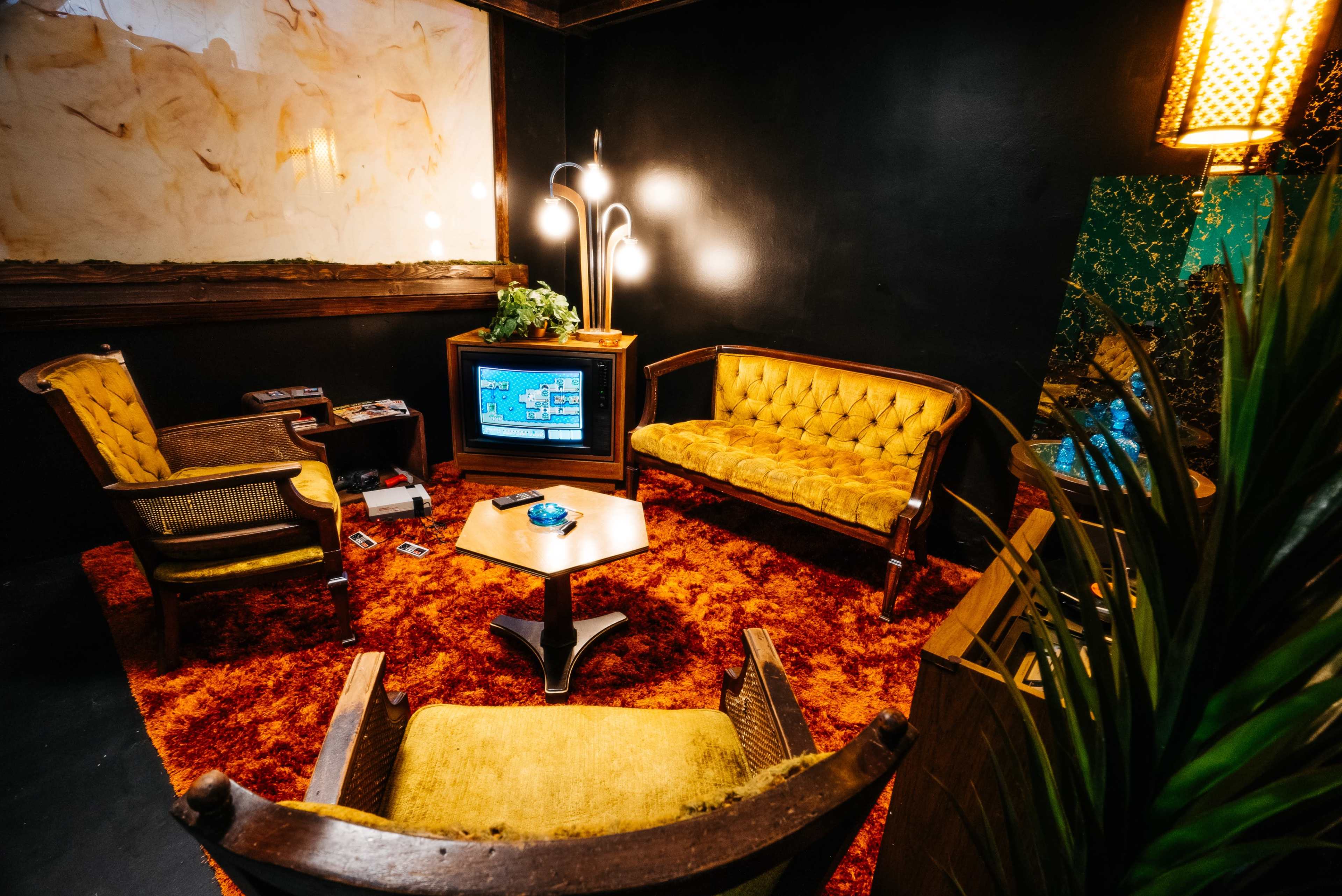 A retro living room with a yellow couch, two armchairs, a small wooden table, and a television set in a dark-walled environment with a patterned carpet.