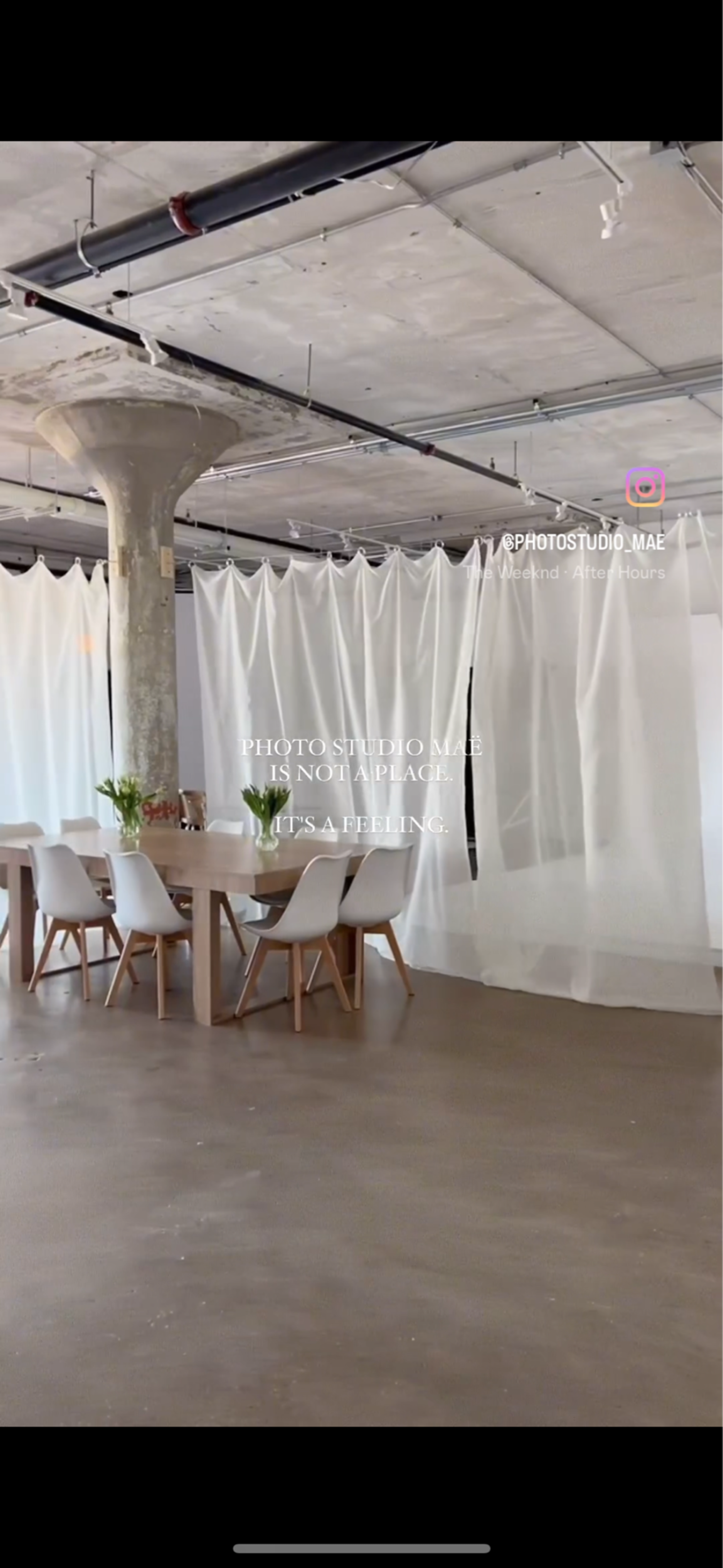 A spacious photo studio featuring a long table with chairs surrounded by sheer white curtains and a concrete pillar.