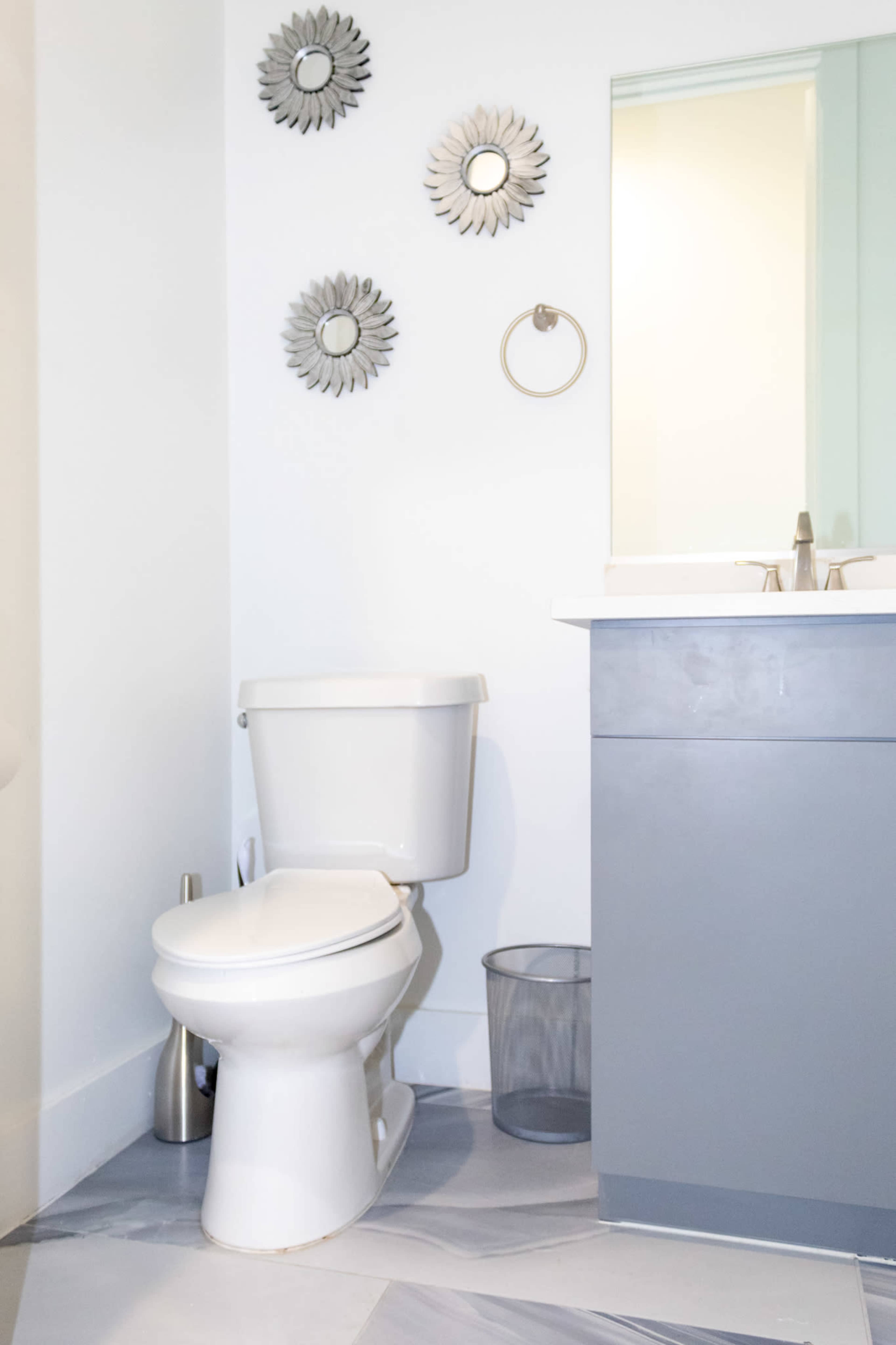 The image shows a bathroom featuring a white toilet, a gray vanity with a sink, and three decorative mirrors mounted on the wall.
