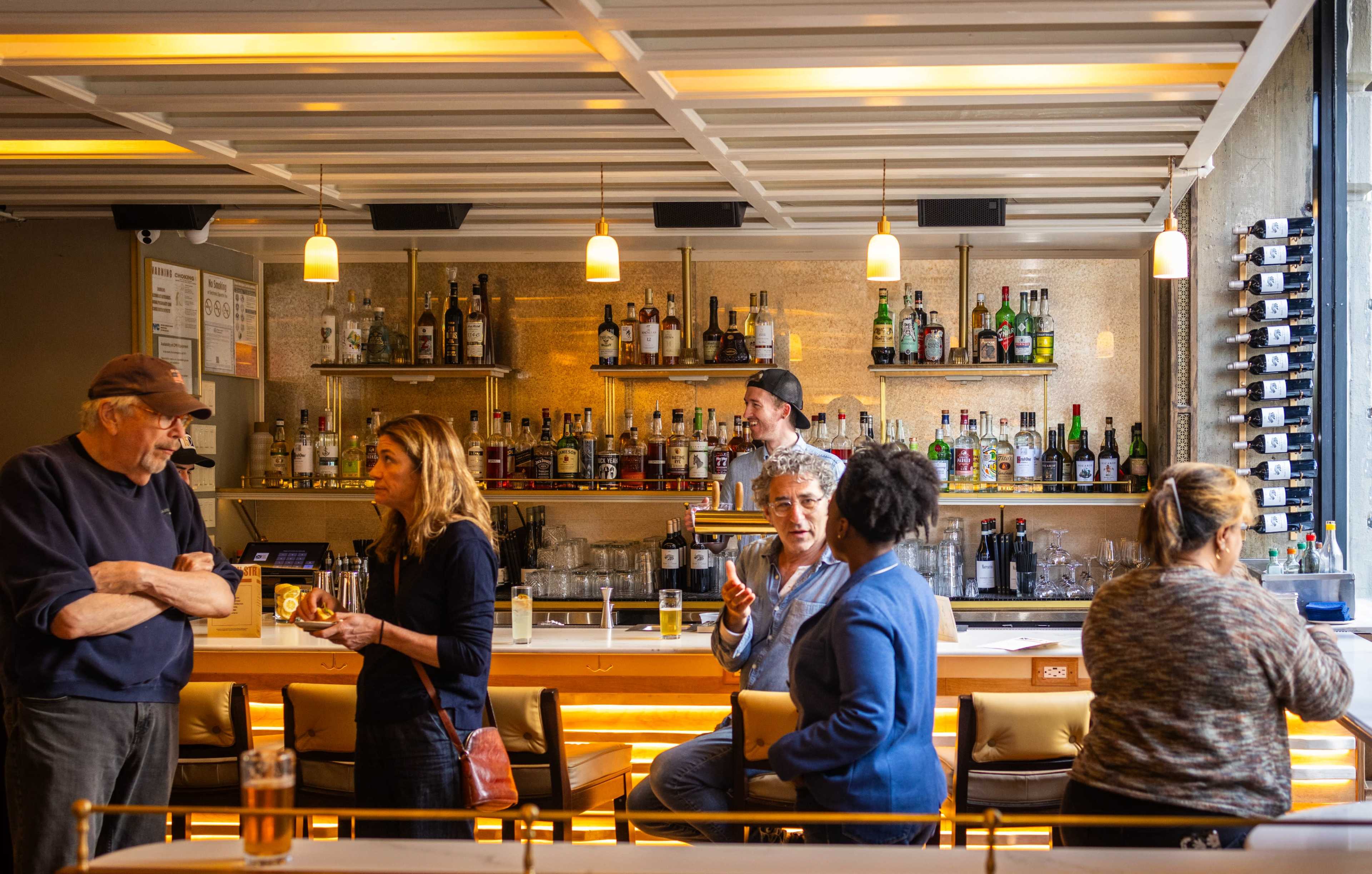A group of people interact in a brightly lit bar, with shelves of liquor bottles displayed behind the counter.