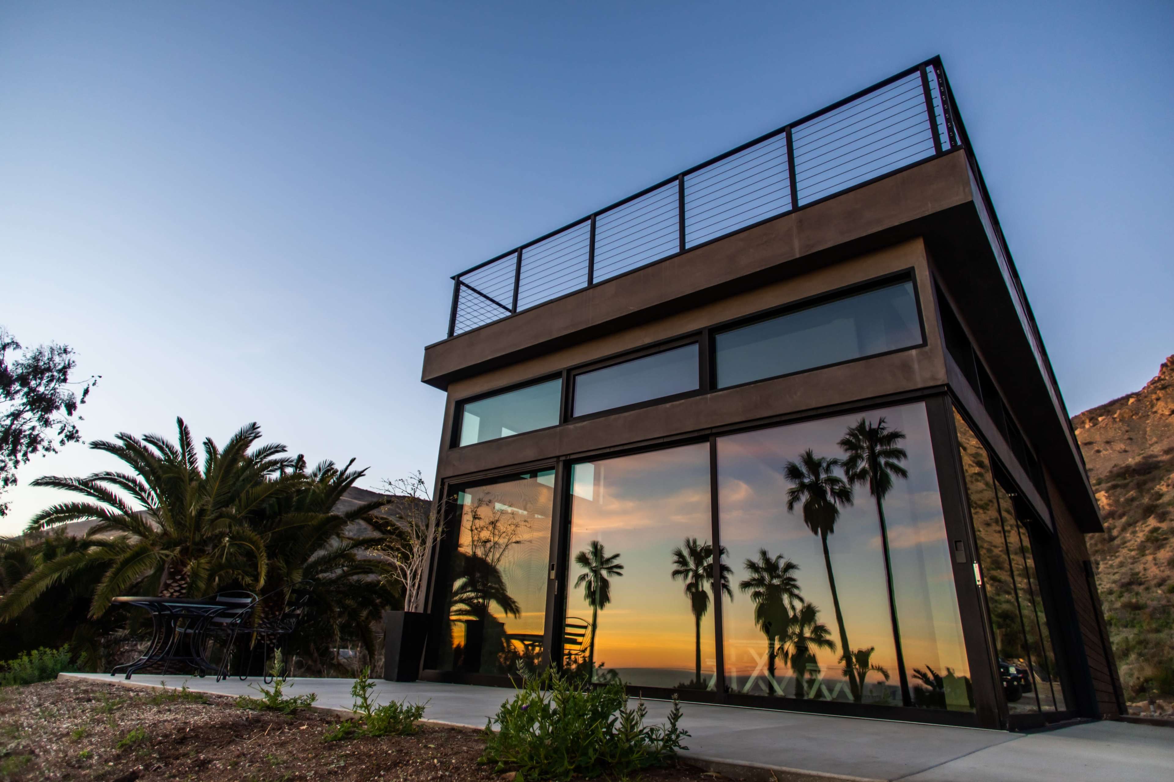 A modern house with large glass windows reflects palm trees and a sunset backdrop.