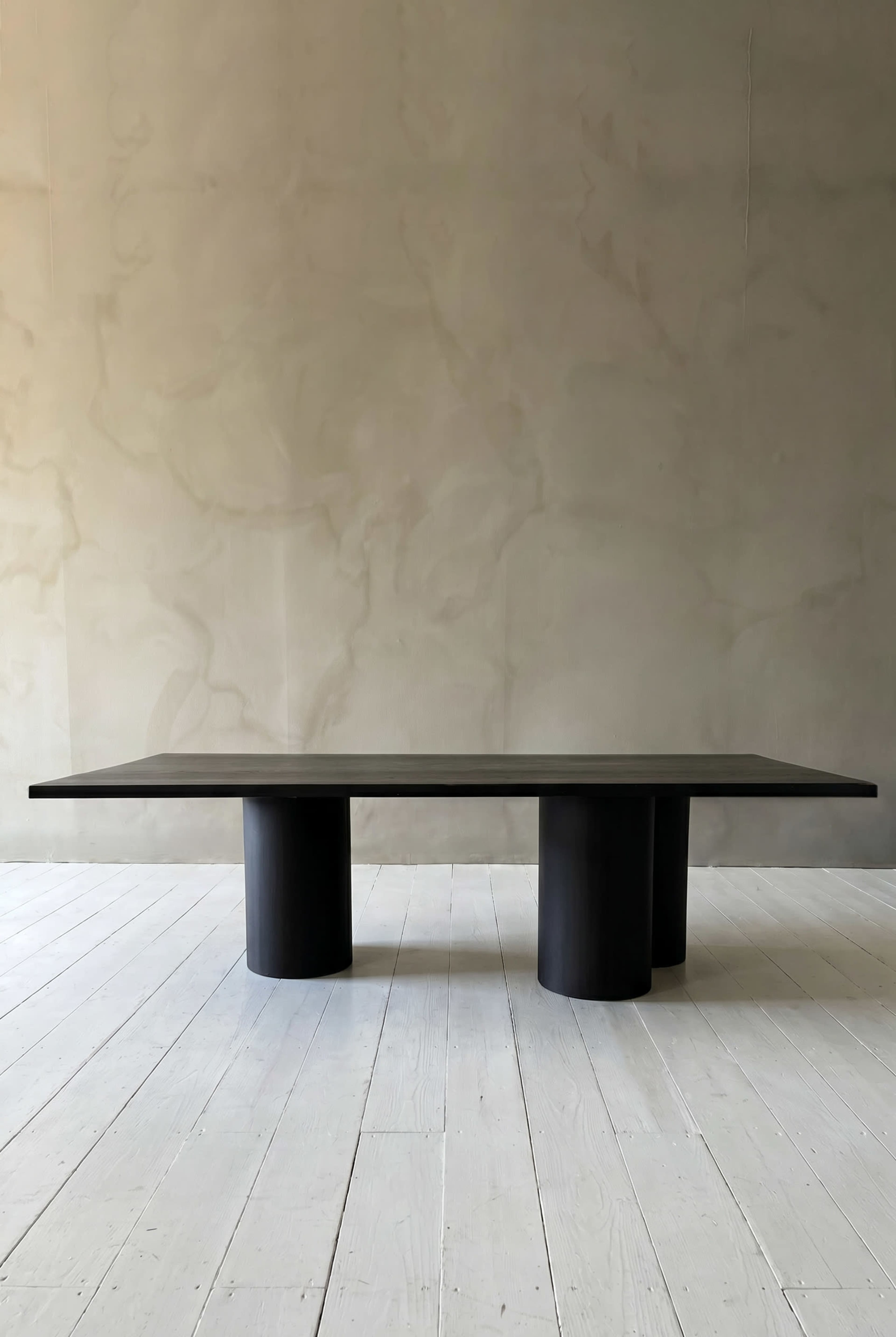 The image features a modern rectangular table with a flat top supported by four cylindrical legs, set against a textured wall and a wooden floor.