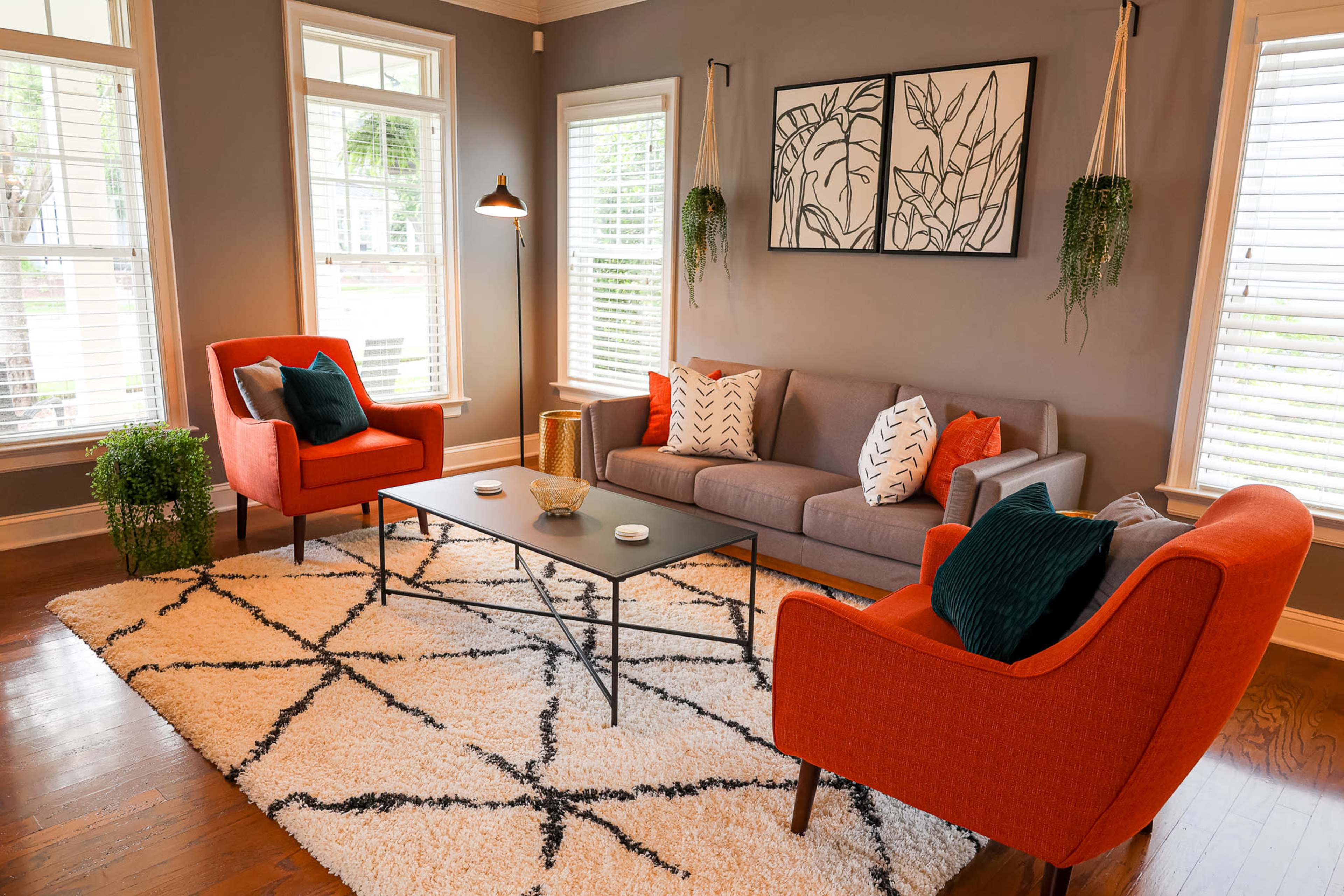 A modern living room with a gray sofa, two orange chairs, a black coffee table, and a patterned rug, surrounded by large windows and decorative wall art.