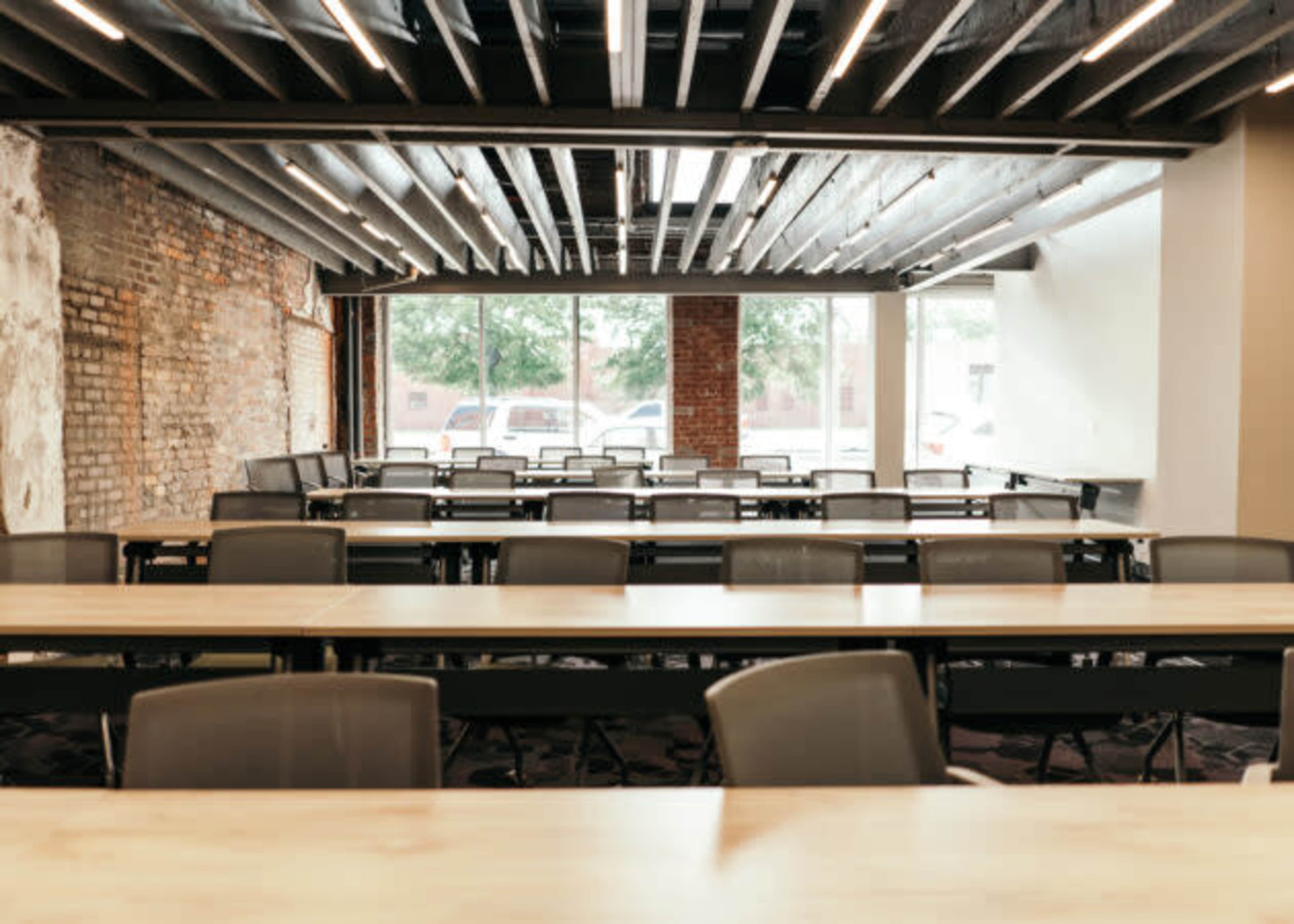 The image shows a spacious, modern meeting room with rows of tables and chairs, featuring exposed brick walls and large windows.