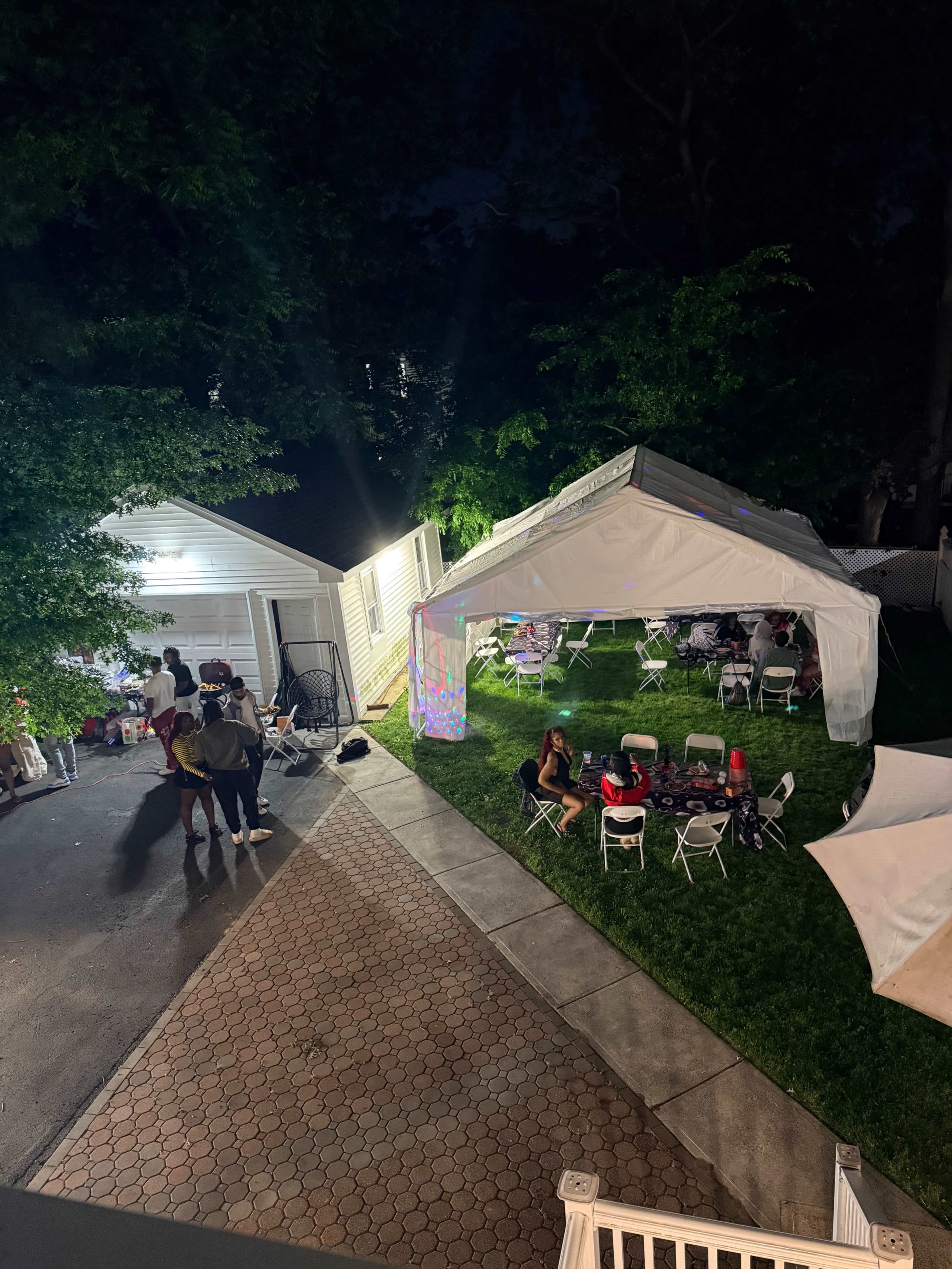 A nighttime backyard scene features a party under a large white tent with chairs set up, surrounded by greenery and a garage.