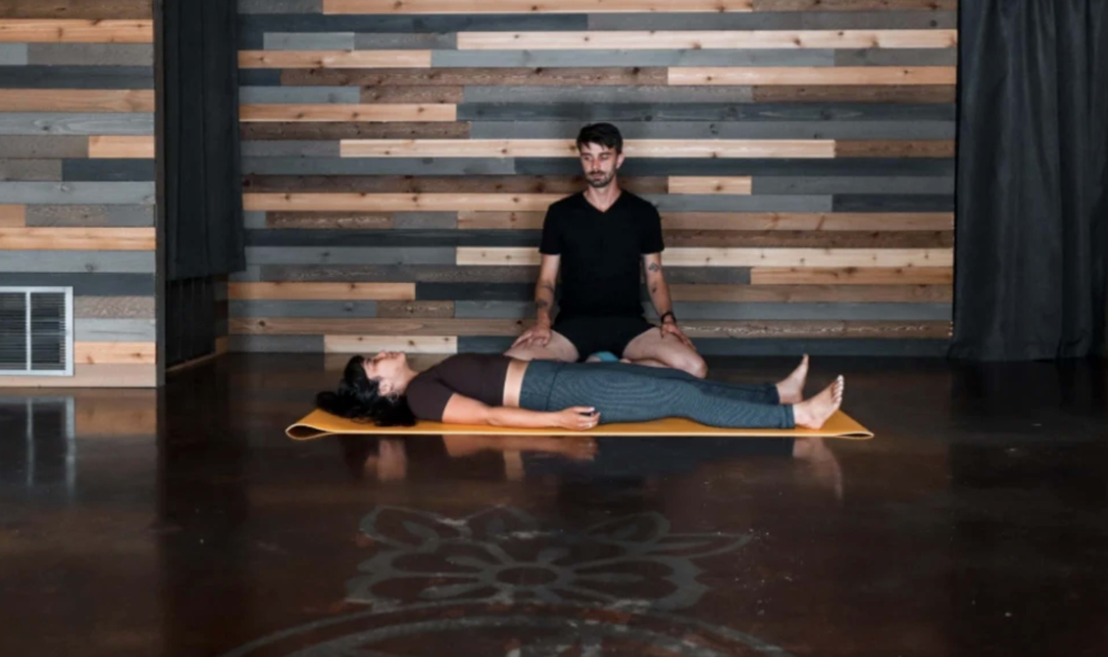 A man sits behind a woman lying on a yoga mat in a room with wooden walls and a polished floor.
