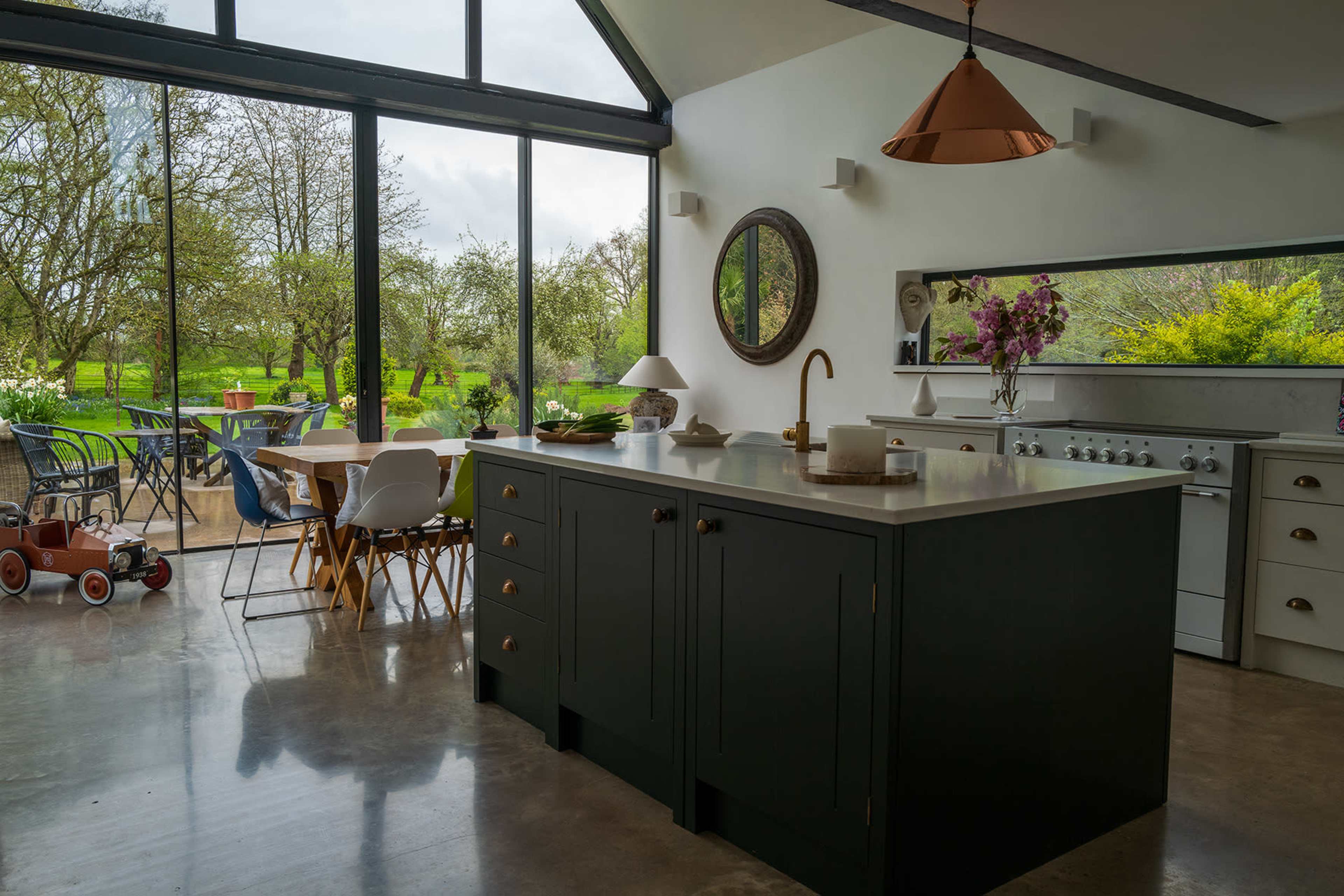 A modern kitchen features a dark island, large windows overlooking a garden, and an assortment of dining chairs and a table.