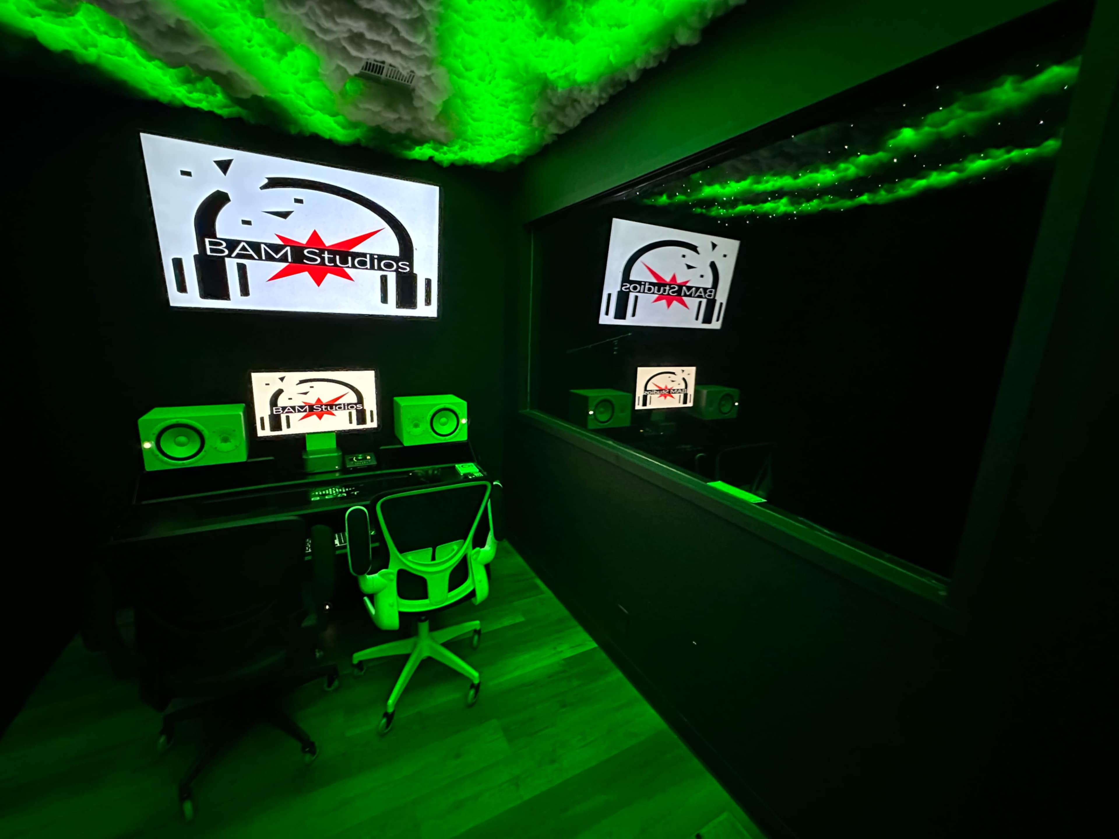 A sound studio equipped with monitors, speakers, and a large display featuring the logo of BAM Studios, with green lighting and a reflective surface.