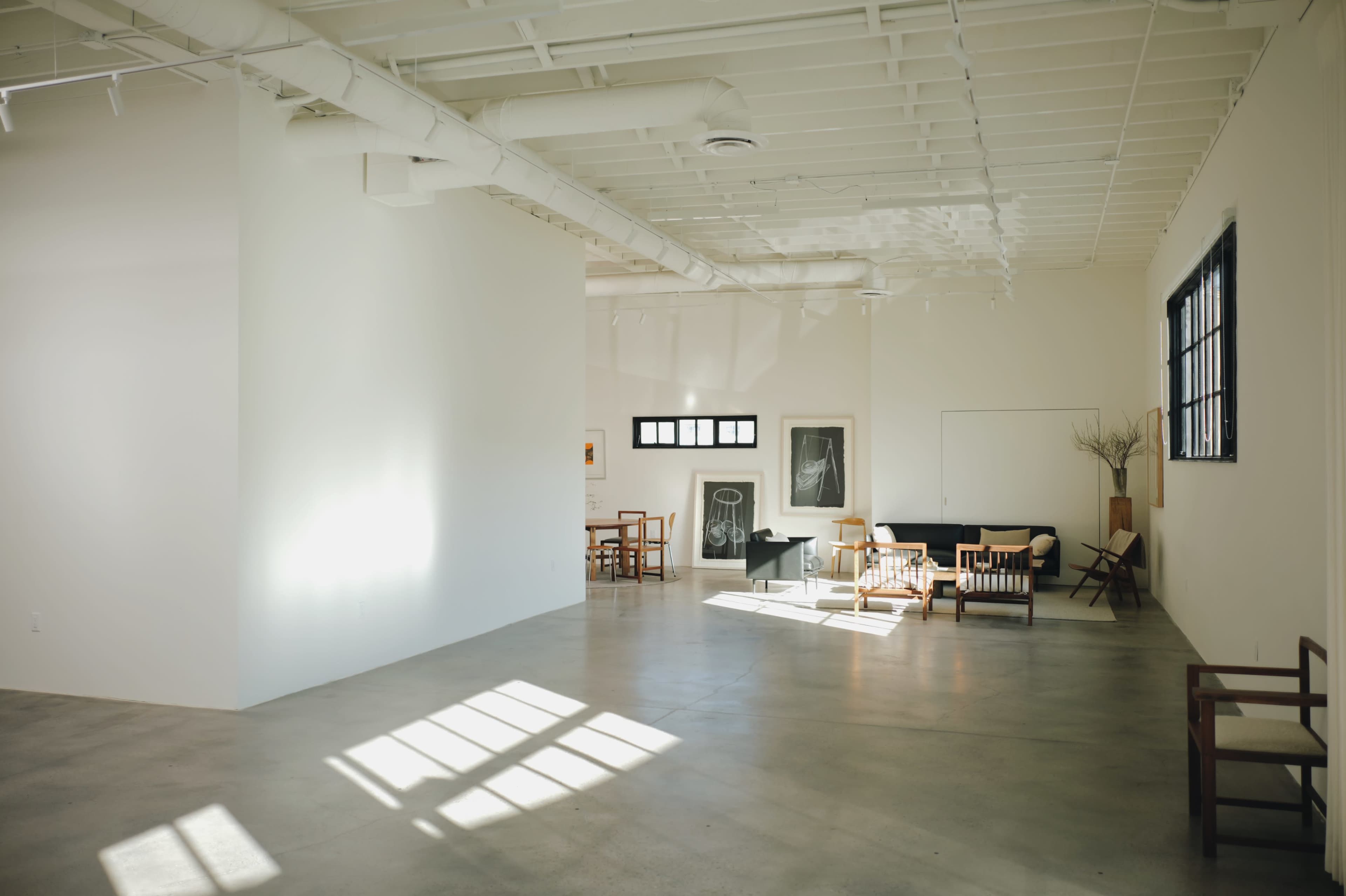 The image shows a spacious, modern interior with a mix of furniture arranged on a concrete floor and large windows allowing natural light to enter.