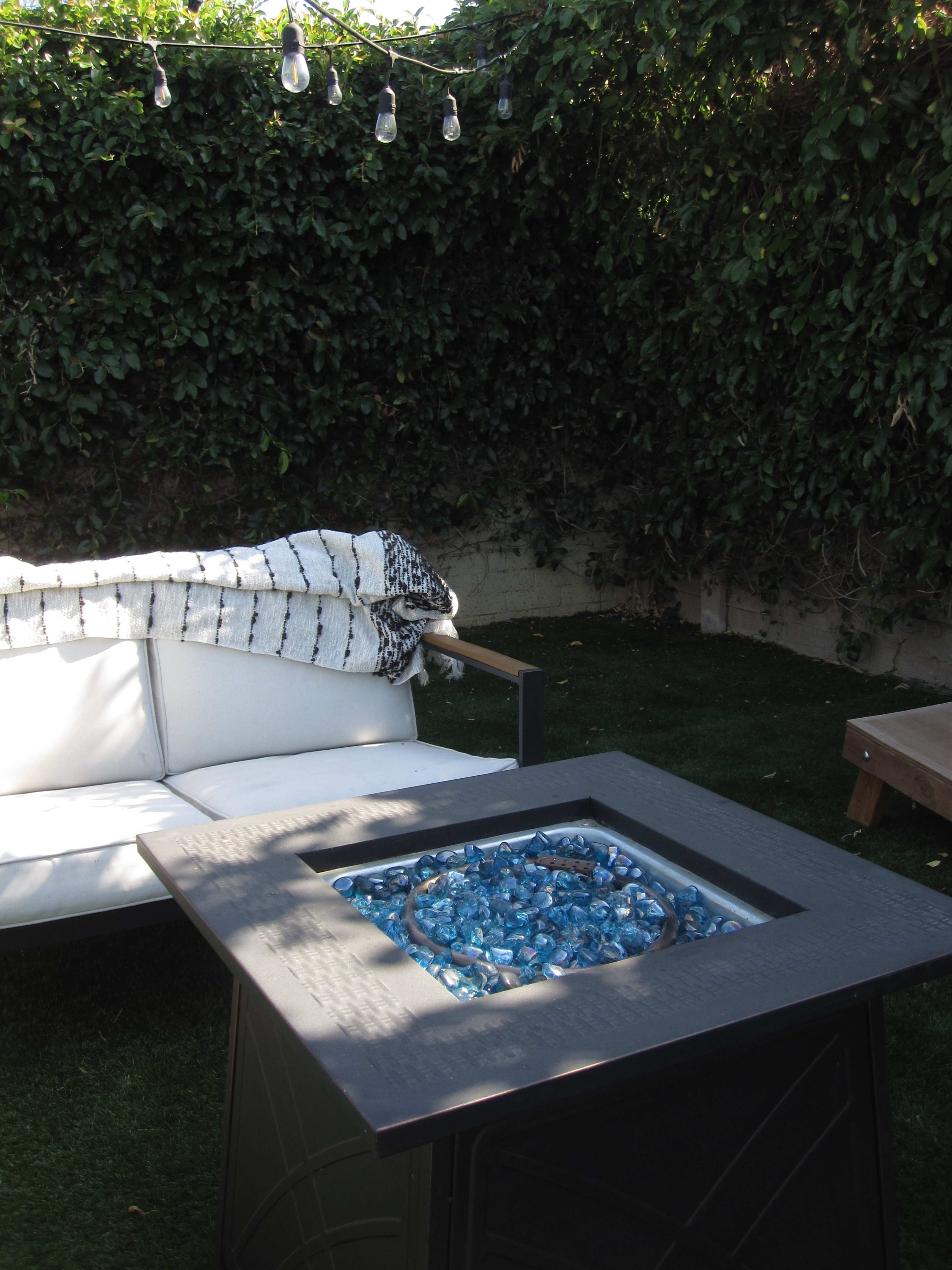 A backyard patio features a modern fire table with blue glass stones, a white couch, and overhead string lights surrounded by lush greenery.